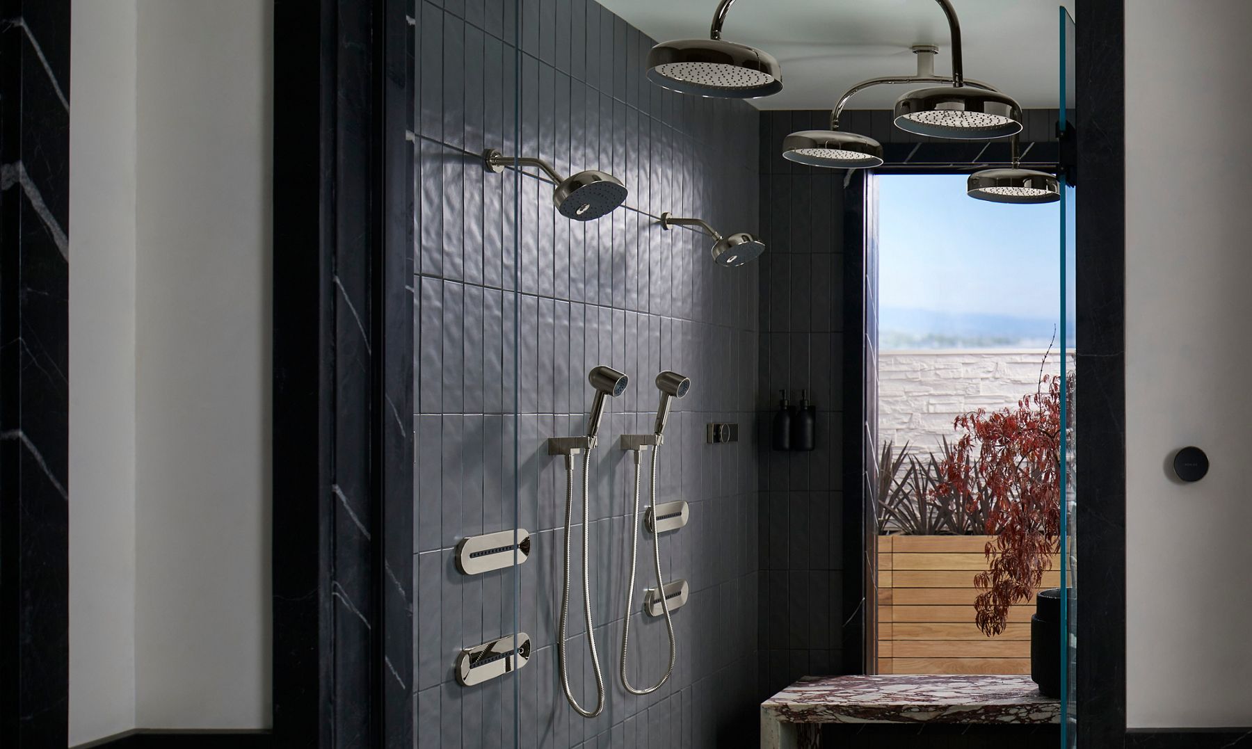 Luxurious modern shower area with dark gray tiled walls and multiple stainless steel showerheads, including both overhead rain showerheads and handheld options mounted on the wall. The sleek, minimalist design features built-in wall controls and a pair of black dispensers. The shower area opens up to an outdoor view through a large window, revealing a wooden deck and surrounding greenery, creating a seamless indoor-outdoor connection.