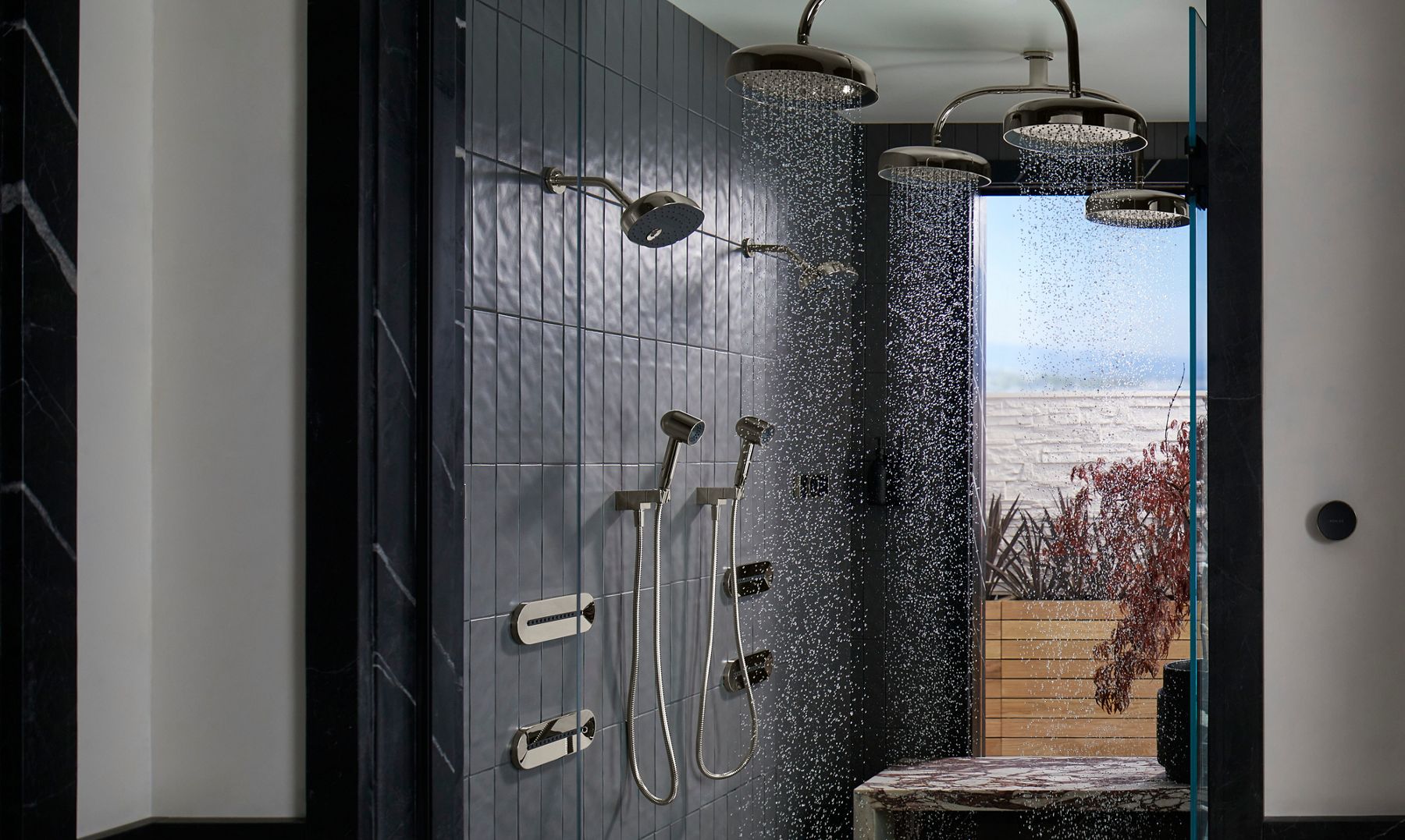Grande image d’une douche moderne avec de grandes pommes de douche à effet de pluie montées au plafond et plusieurs jets corporels latéraux. L’eau coule, produisant un jet dynamique de gouttelettes d’eau en mouvement. La vue à travers la paroi vitrée révèle une scène de bord de mer, ajoutant une toile de fond sereine à l’installation de la douche haut de gamme.