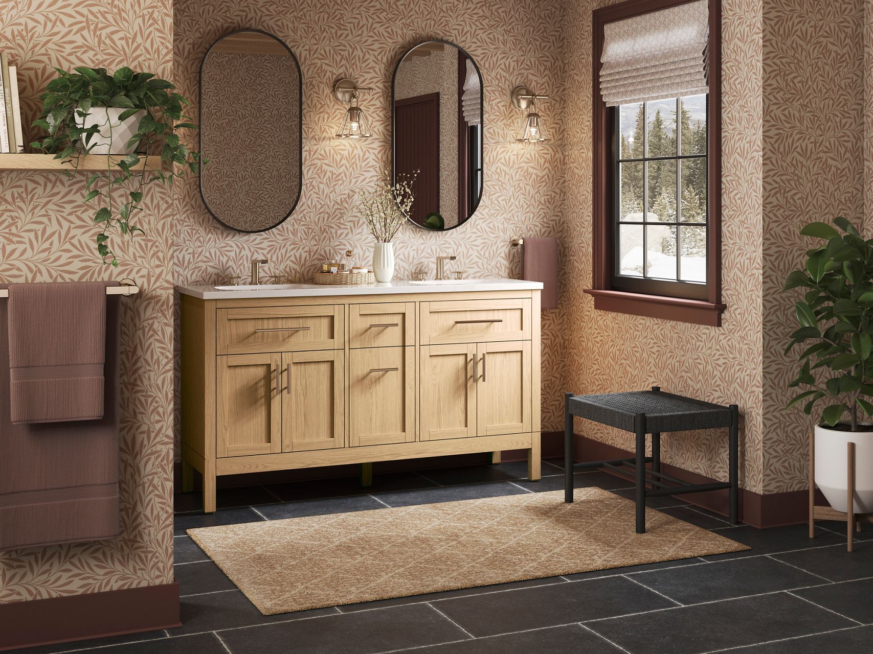 Modern bathroom with a light wood double vanity featuring sleek hardware and a white countertop. Above the vanity, there are two oval mirrors with black frames, illuminated by industrial-style wall sconces. The wall behind the vanity is covered in a beige and white botanical patterned wallpaper, adding texture and warmth to the space. A small vase with delicate branches and a tray with toiletries are placed on the countertop, enhancing the clean and minimalist design. The dark floor tiles contrast with the light tones of the vanity and wallpaper, creating a balanced and inviting atmosphere.
