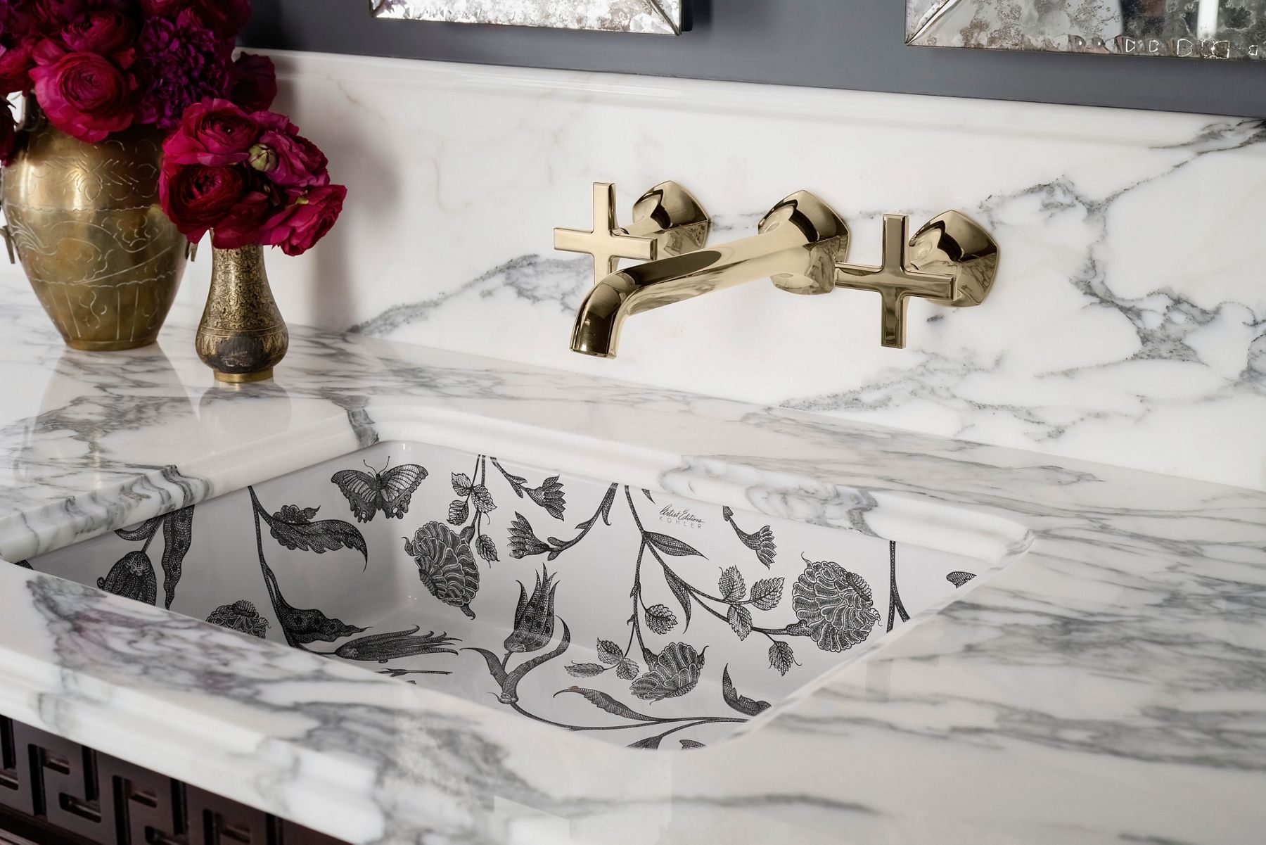 Gold wall-mounted gold faucet with cross handles above a marble counter and sink with black floral design.