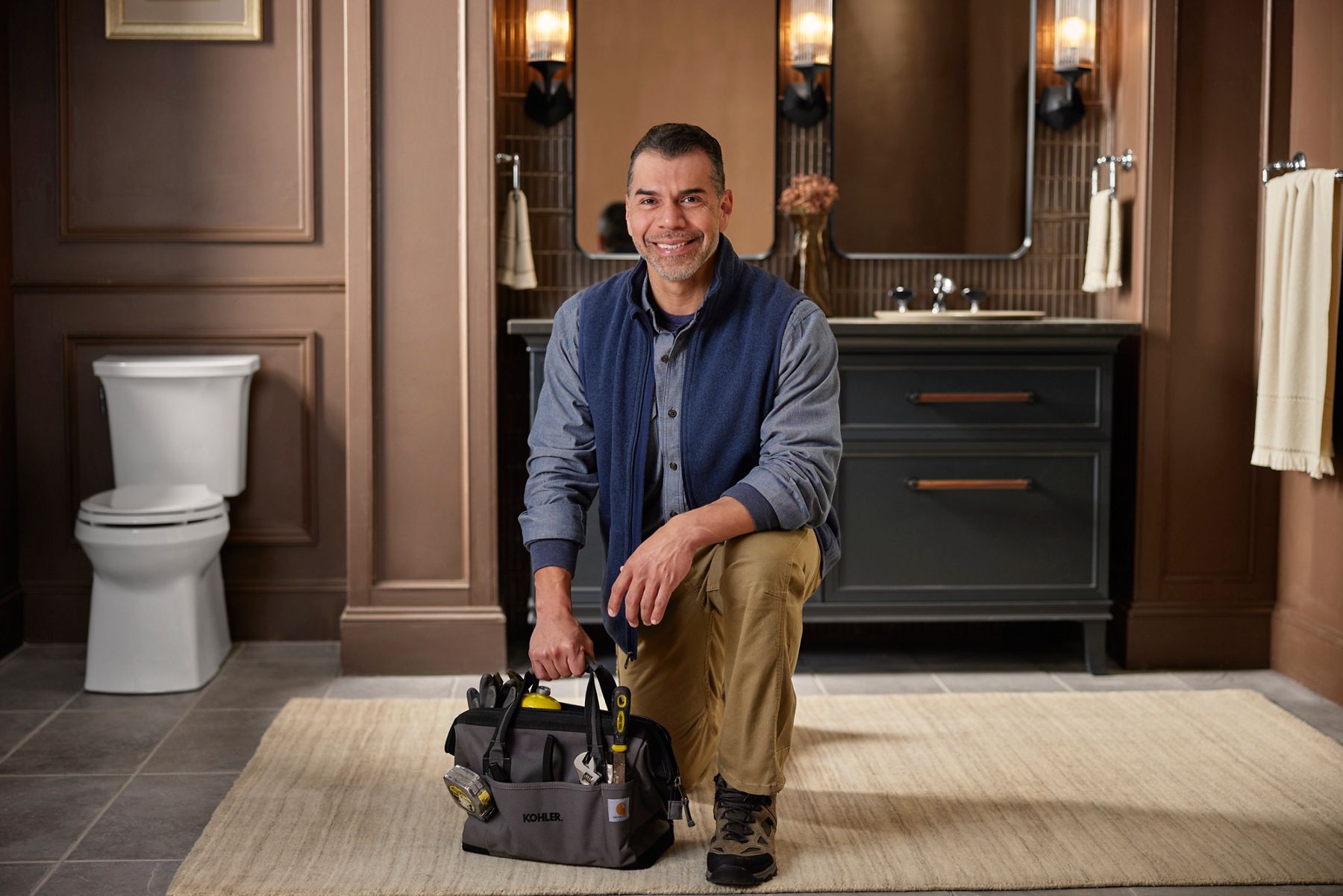 Smiling Kohler installer kneeling on the floor, holding a tool bag with Kohler branding, set in a modern bathroom with dark cabinetry and a large mirror.