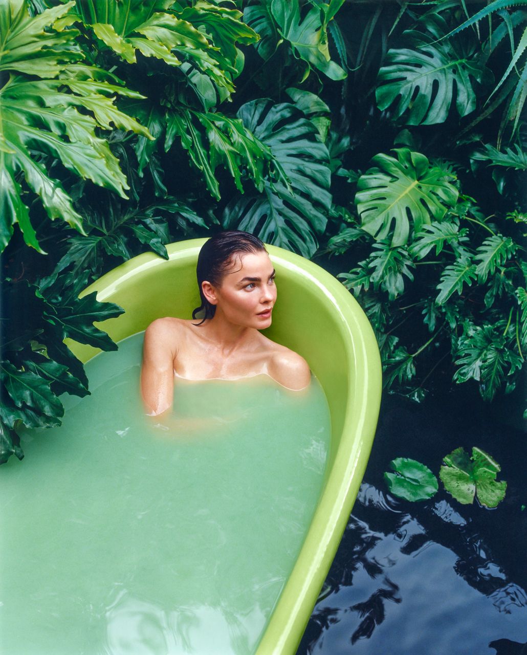 A slightly above portrait shot of a woman in a bath tub filled with water. The bathtub is a bright lime green and positioned in a garden surrounded by greenery and a pond.