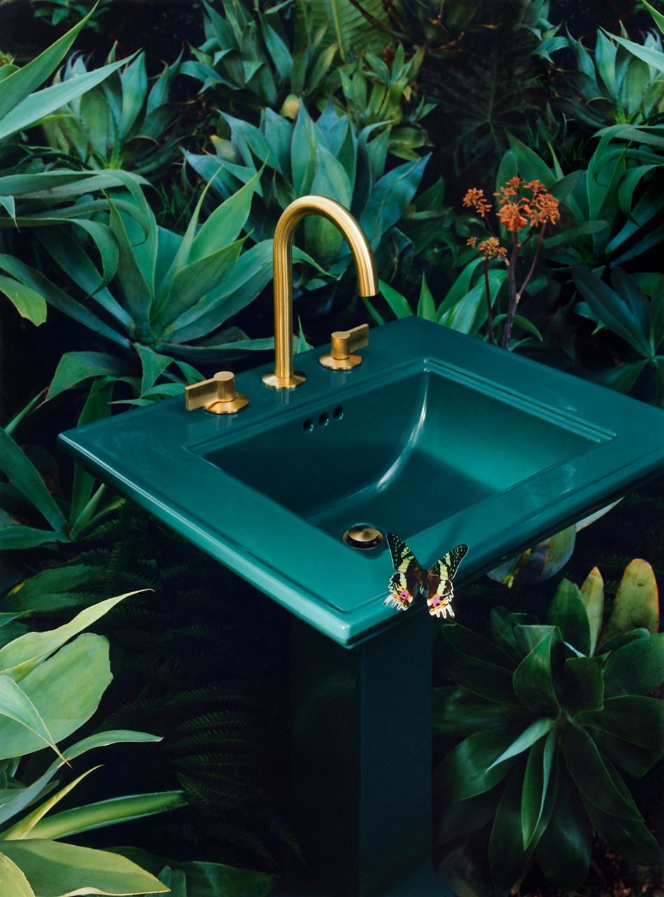 A portrait image of a teal standing bath sink positioned in the middle of a garden. The sink is designed with a gold faucet, gold handles, and a gold drainer. There is a yellow butterfly sitting at the edge of the sink.