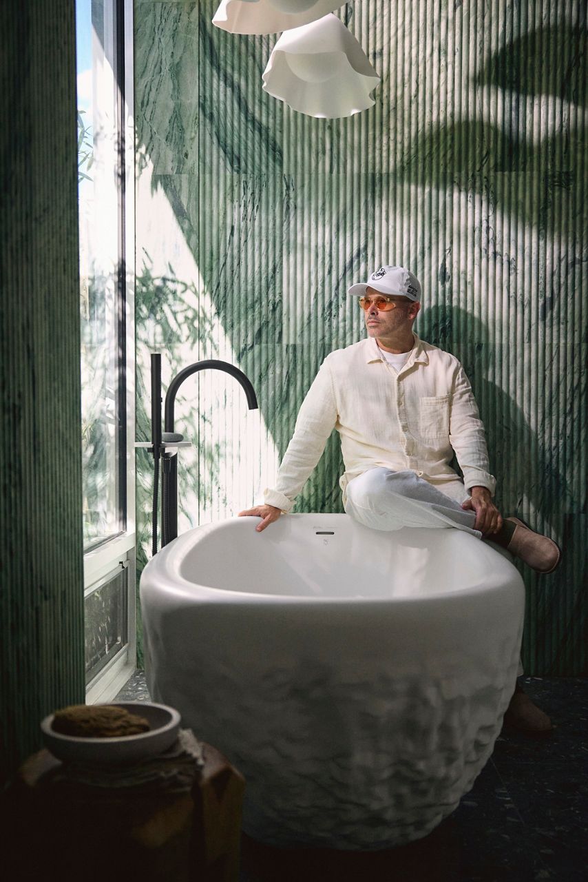 Artist Daniel Arsham sits on the edge of a white abstract bathtub against a green textured marble wall.