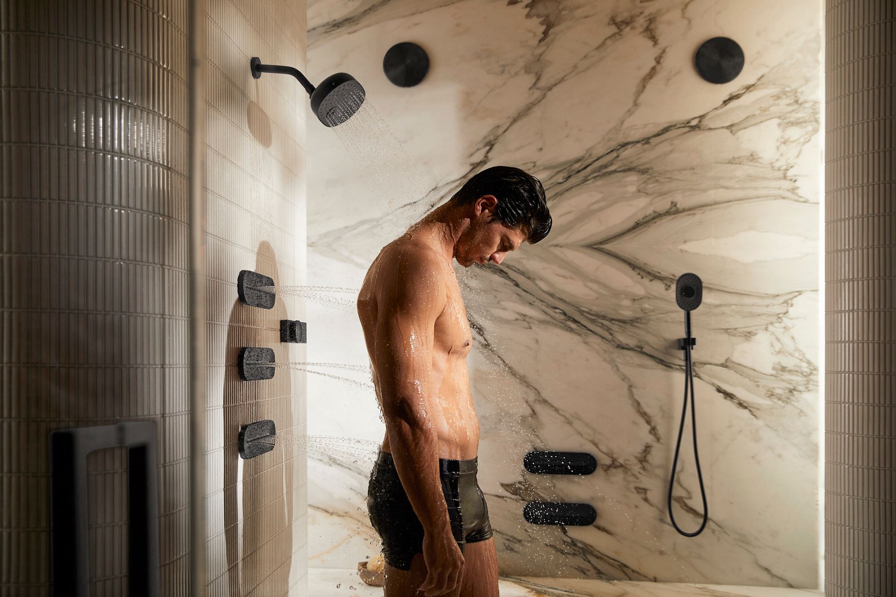 A man standing in a modern shower with water spraying from multiple showerheads, including an overhead and several wall-mounted nozzles. The shower is set against a marble wall backdrop, and the man is facing downwards, enjoying the water.