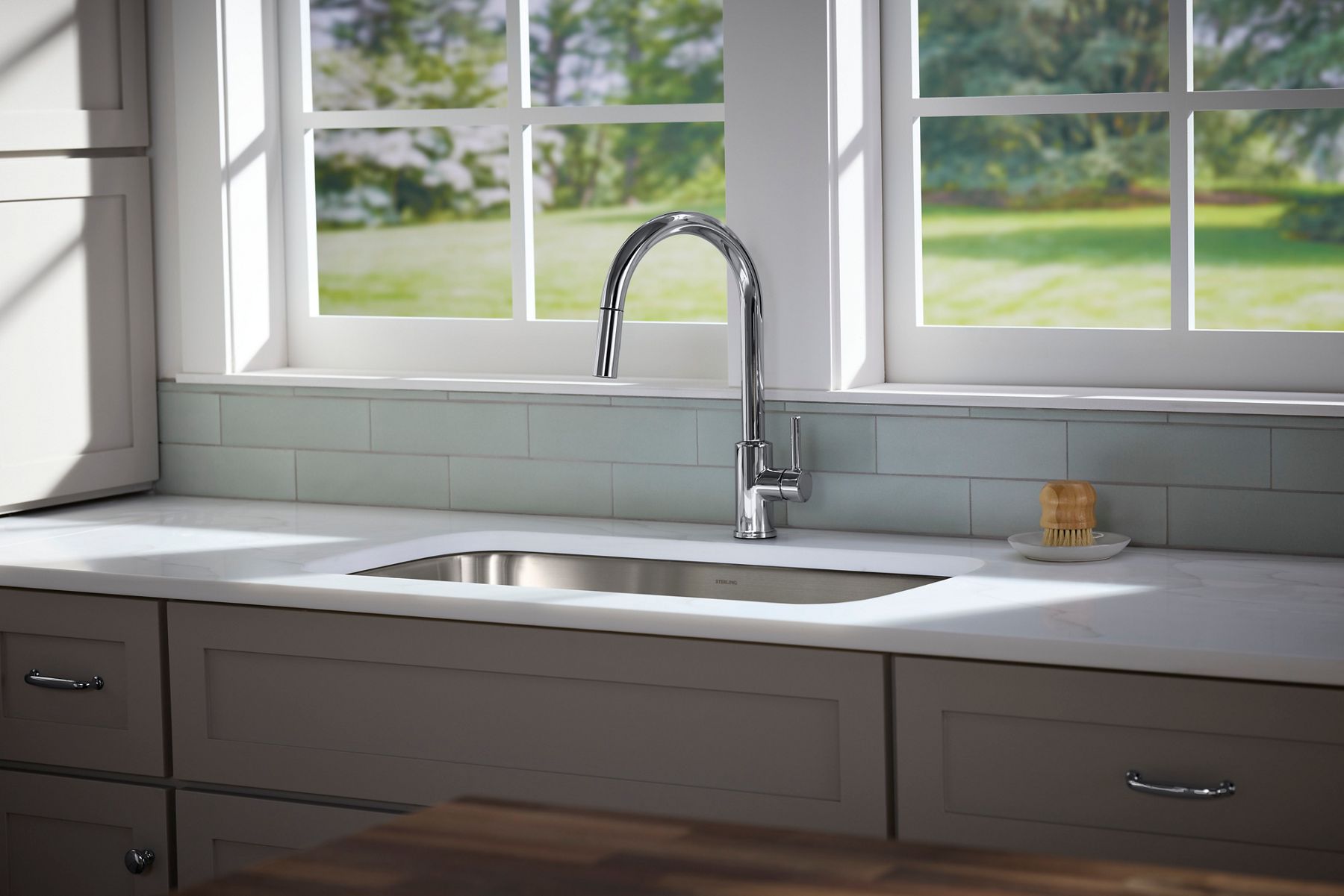 Kitchen sink with a polished chrome pull-down faucet centered beneath large multi-pane windows, set against a light tile backsplash and white countertop in a bright, neutral kitchen.