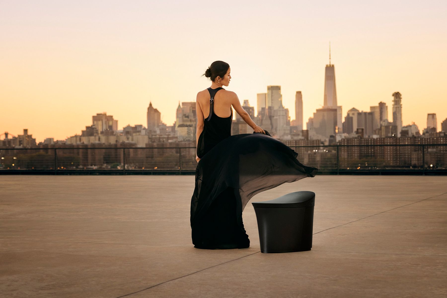Laura Kim stands in a flowing black chiffon dress, accentuating the curved design of the Veil smart toilet against the New York City skyline.