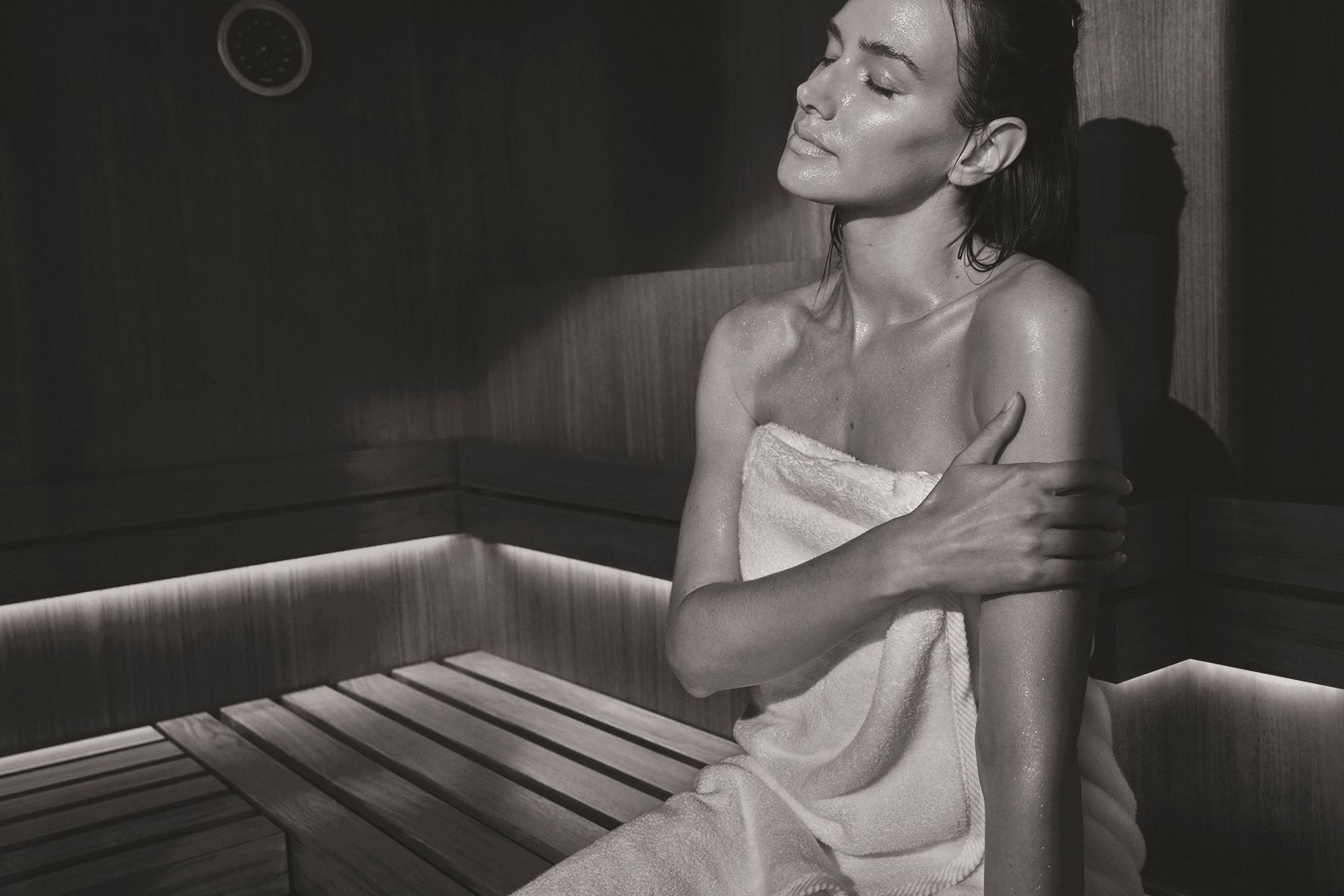 Woman with wet hair wrapped in a towel, gently touching her shoulder, eyes closed in relaxation.
