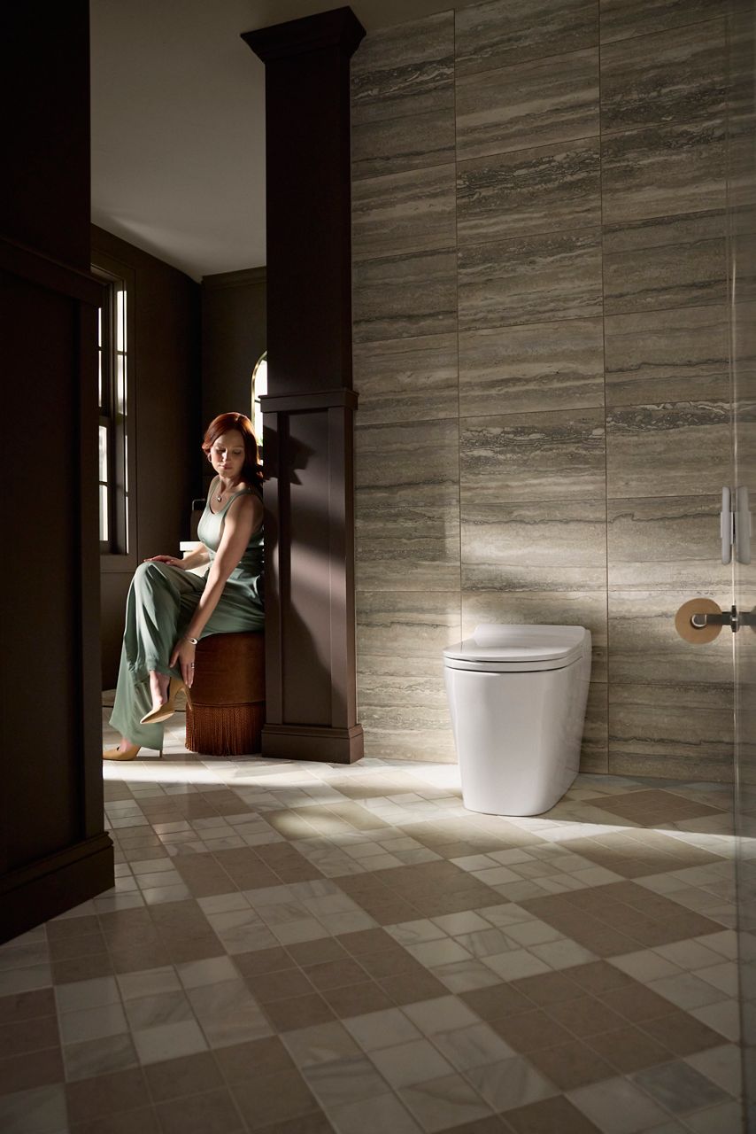 A softly lit modern bathroom with large stone tile walls and a patterned tile floor. A woman sits on a bench beside a dark column, looking outward the camera while a sleek white toilet is positioned against the wall to the right.