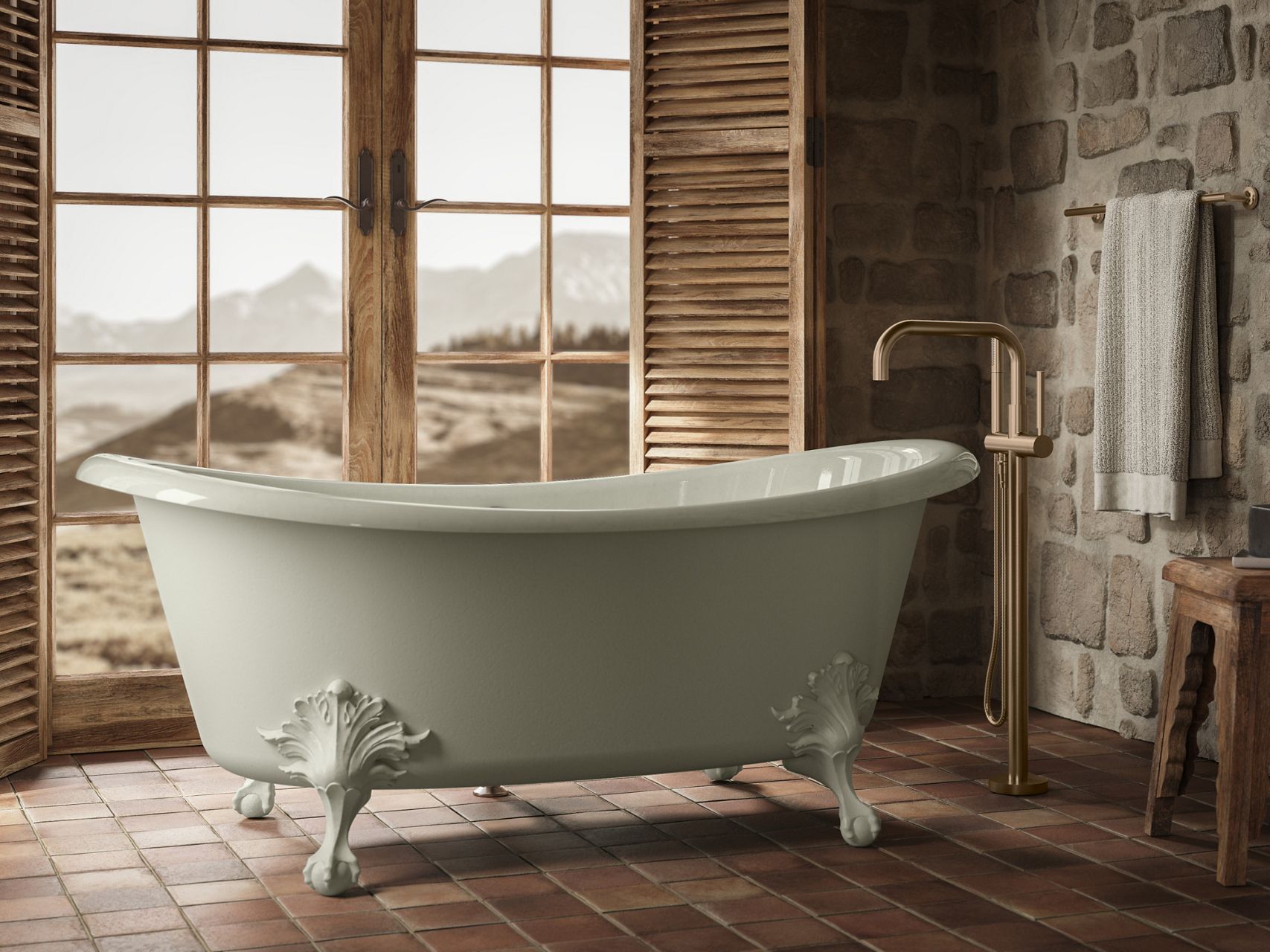 White freestanding clawfoot bathtub in a rustic bathroom with stone walls, terracotta flooring, and large wood-framed windows overlooking a desert landscape.
