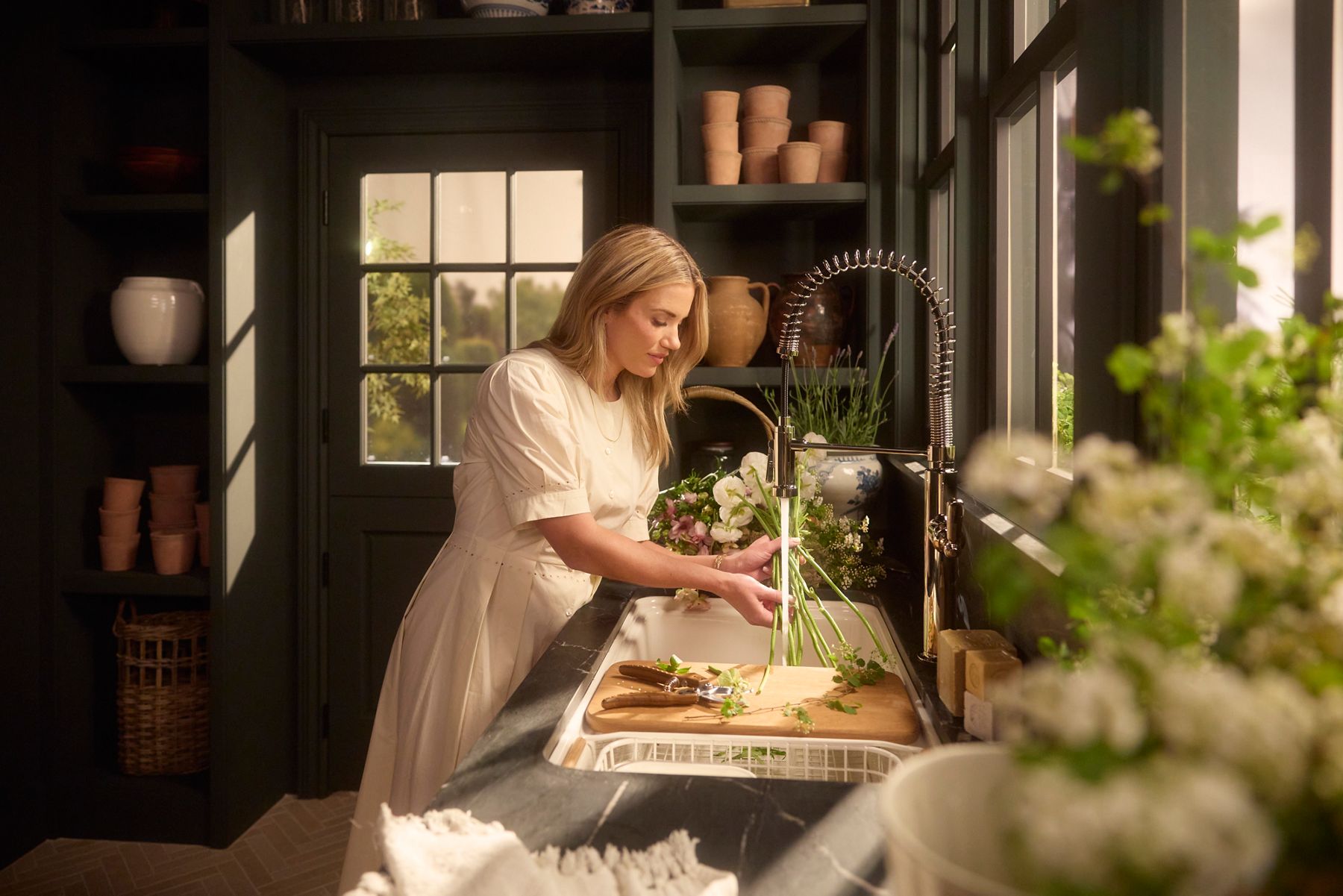 Shea McGee rinses flower stems at a farmhouse-style kitchen sink, with a cutting board, scissors, and fresh greenery on the counter, warm sunlight filling the room.