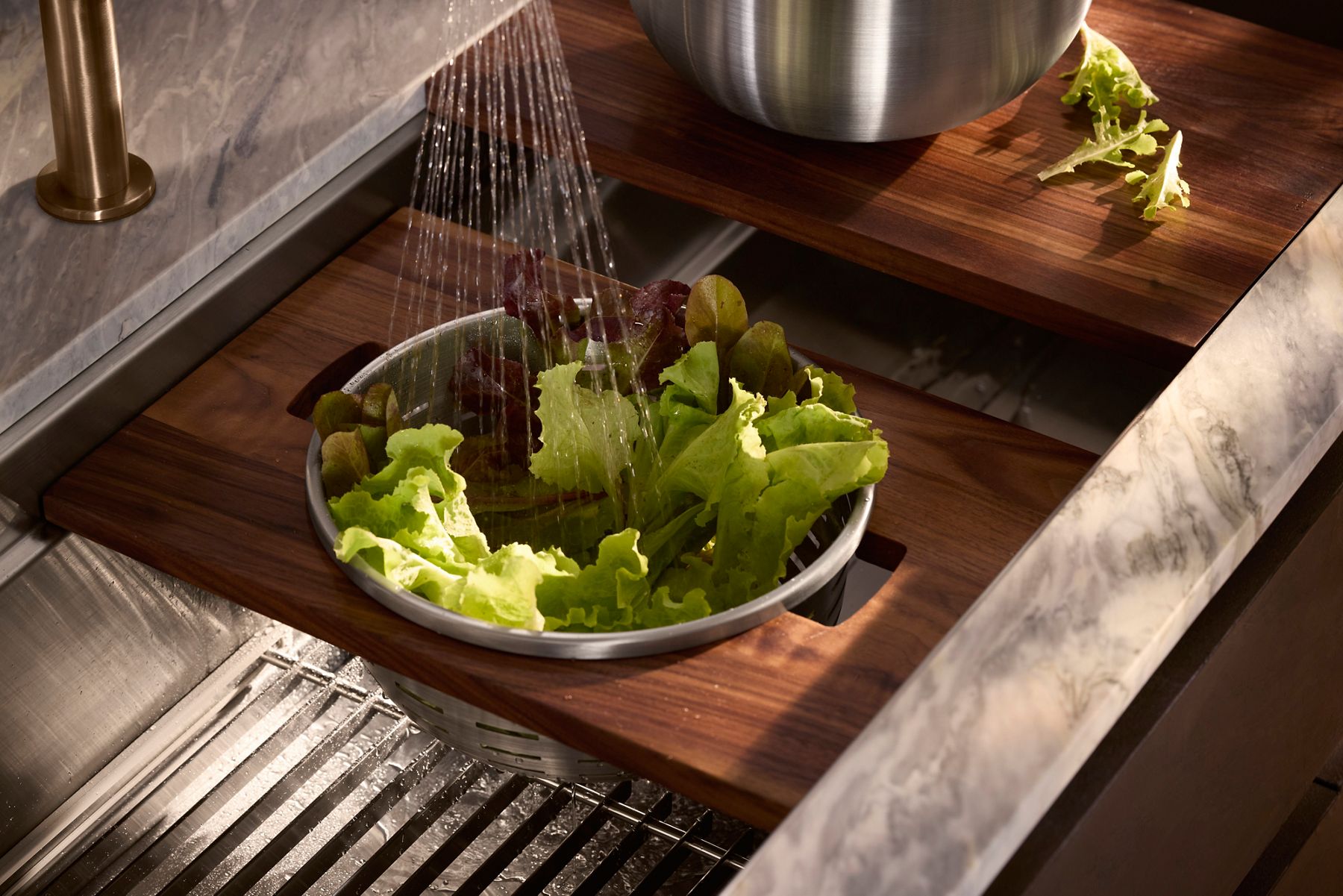 Lettuce being rinsed in a colander inside a KOHLER kitchen sink with a cutting board.