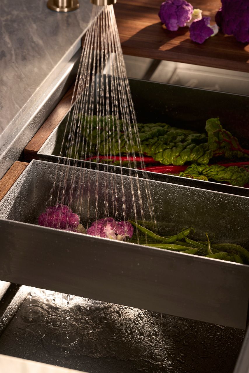 Water rinsing vegetables in stainless steel trays inside a KOHLER kitchen sink.