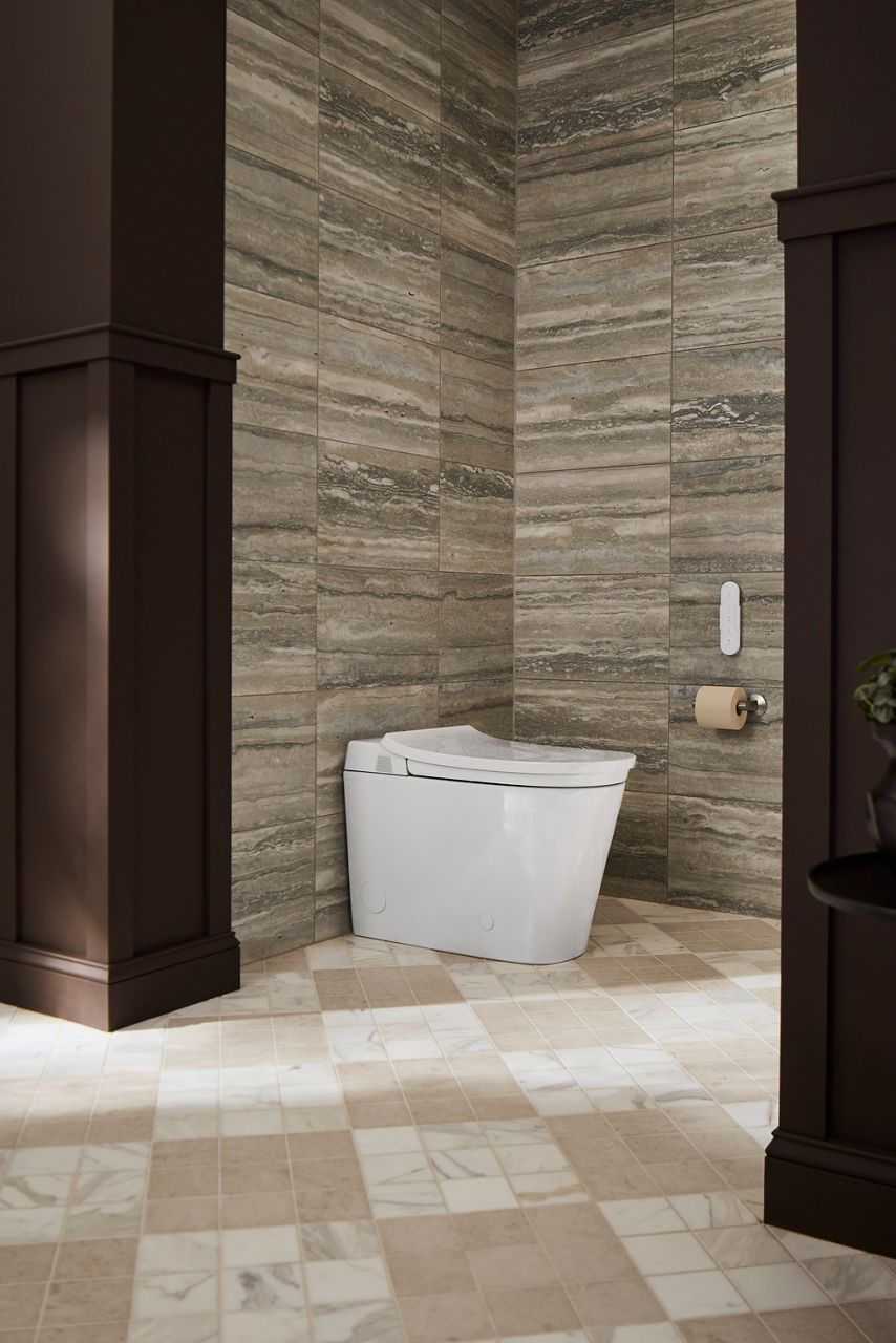 White smart toilet in a sunny bathroom with gray stone walls, cream tiled flooring, and a dark wood entryway.