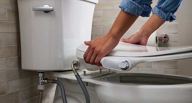 Hands installing a white bidet toilet seat onto a toilet in a bathroom with exposed water supply connection visible.