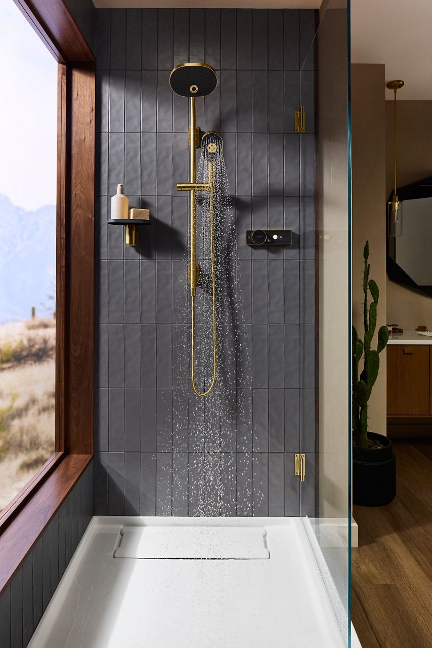 A close up of Kohler shower system featuring a rectangular rainhead and matching handheld showerhead mounted on a slide bar against a tiled wall. Water flows from the handheld showerhead, showcasing the showering components.