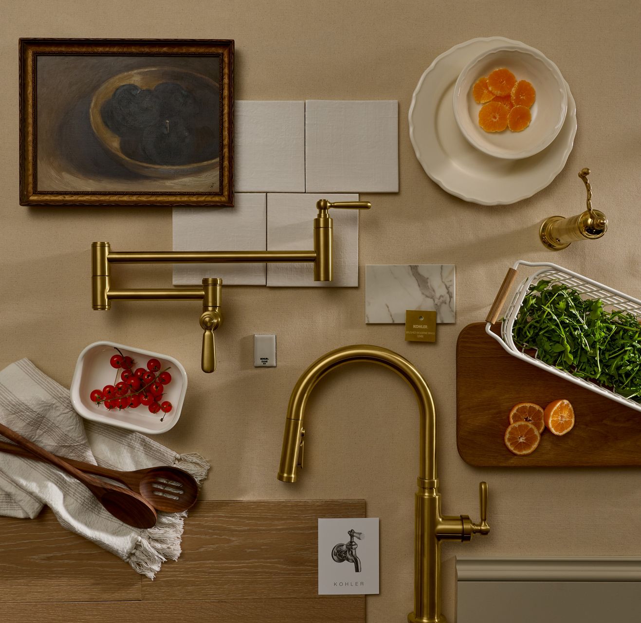 Flat lay of the Shea McGee x Kohler kitchen collection featuring brushed brass kitchen faucets, hardware, and fixtures styled alongside marble tile samples, framed artwork, fresh ingredients, and wooden kitchen utensils on a textured neutral surface.