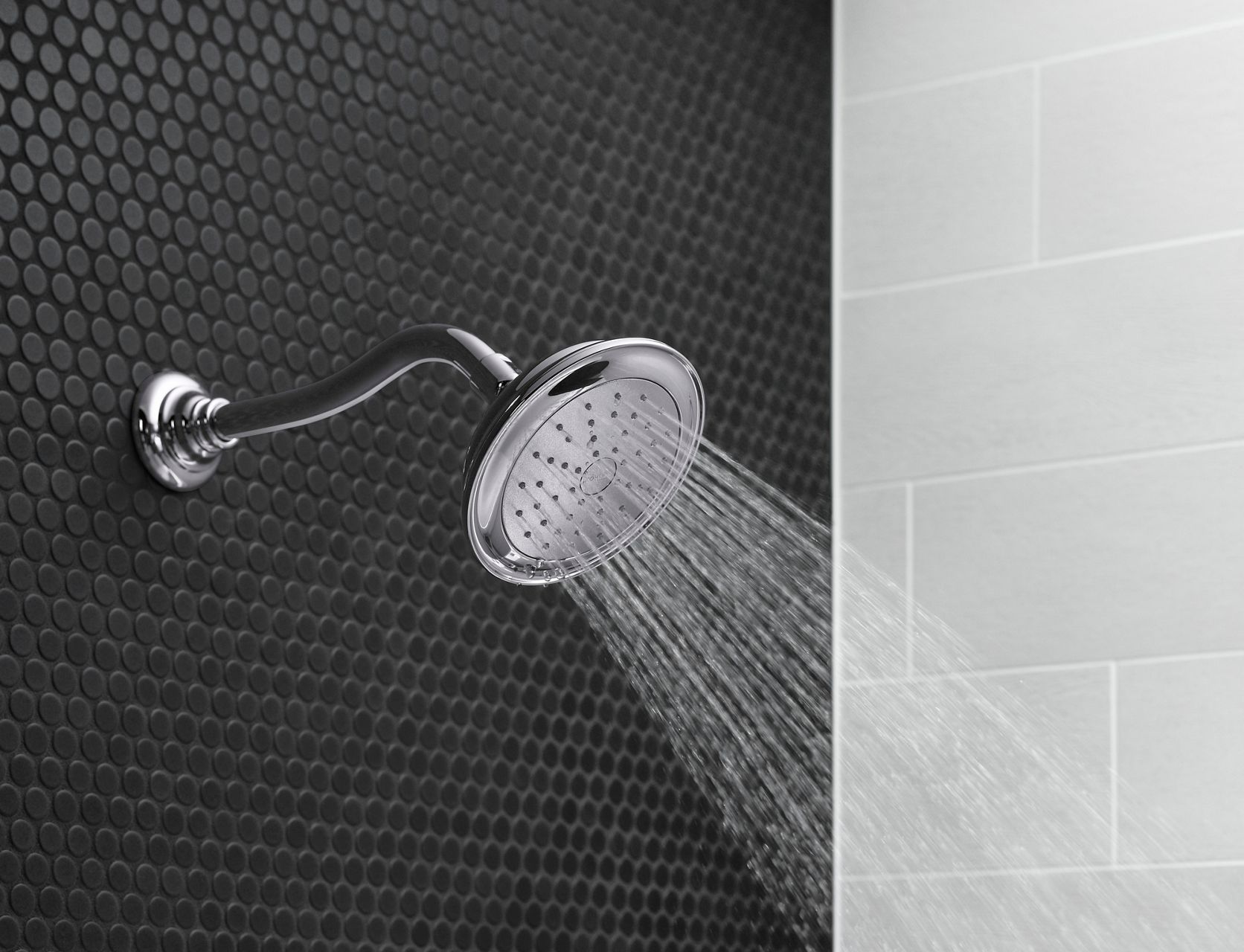 Chrome single-function wall-mounted showerhead with a curved shower arm spraying a steady stream of water against black penny tile and light gray subway tile walls.