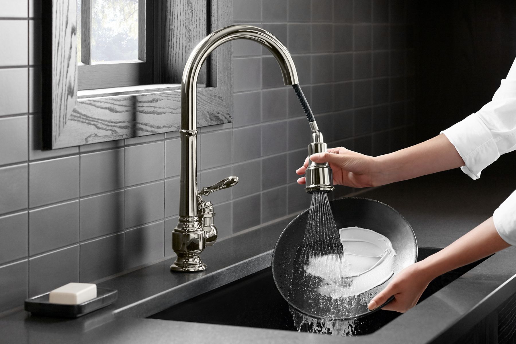 Two hands holding a plate and the spout of a faucet over a sink as water from the sweep spray function washes the plate clean.