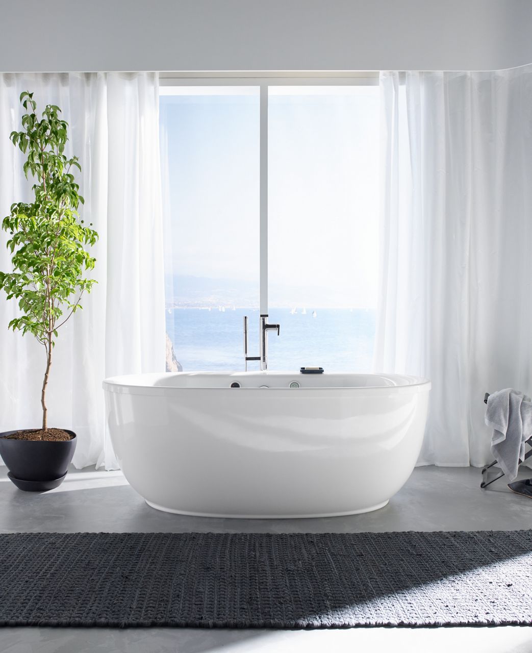 A potted tree stands next to a white KOHLER Sunstruck oval BubbleMassage Air bath with Composed freestanding bath filler and handshower.