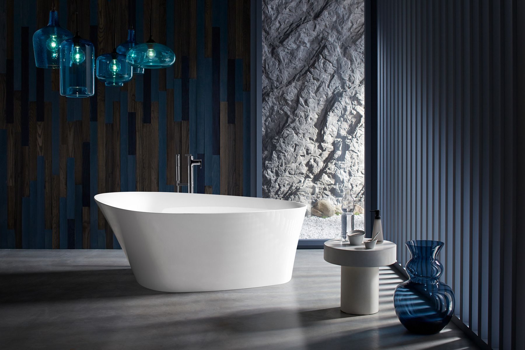 Pendant lights with blue glass shades hangs above a white KOHLER Veil freestanding bath.