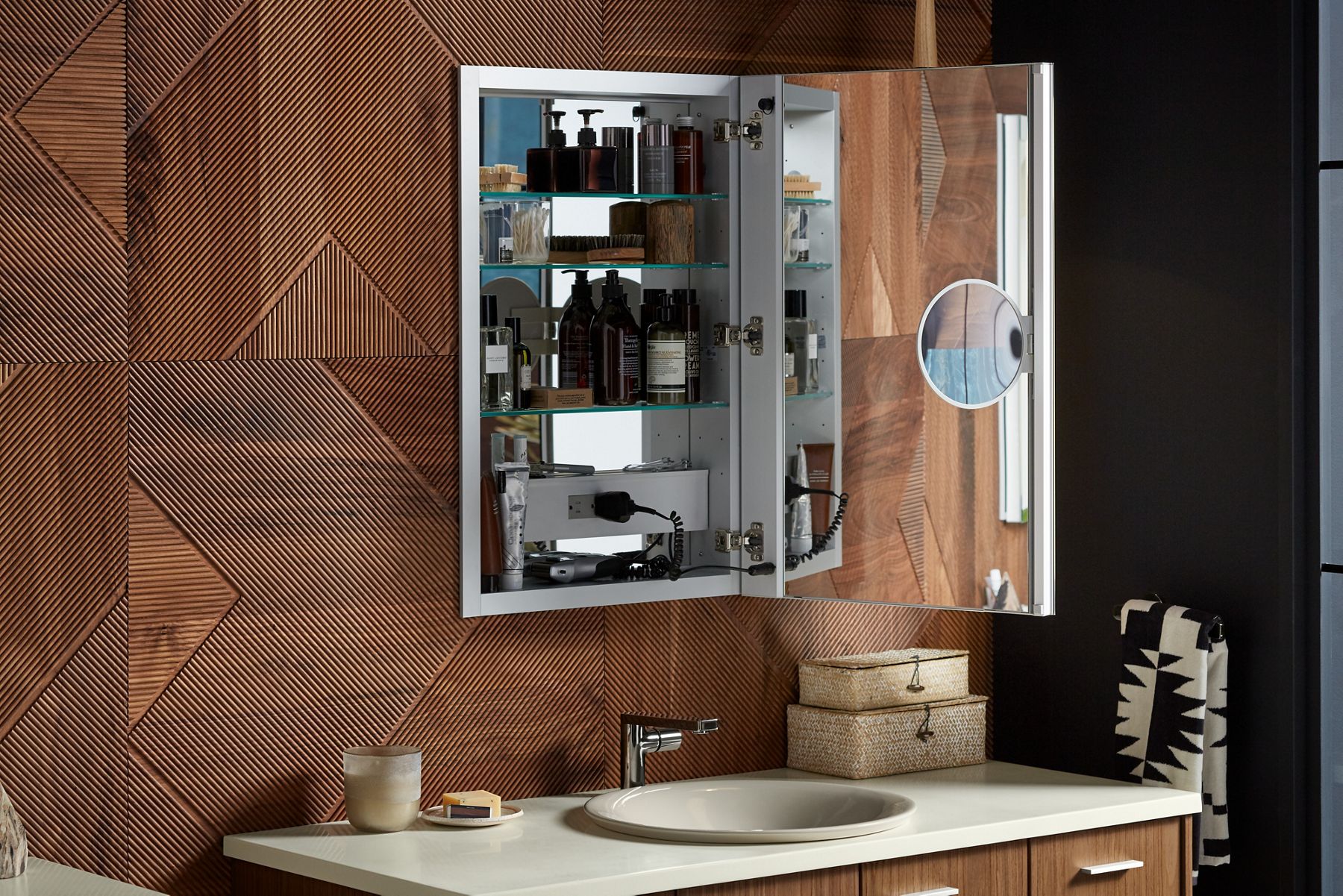 An open KOHLER medicine cabinet with a mirrored interior and adjustable magnifying mirror hangs above a wood, white-topped vanity with a drop-in sink.