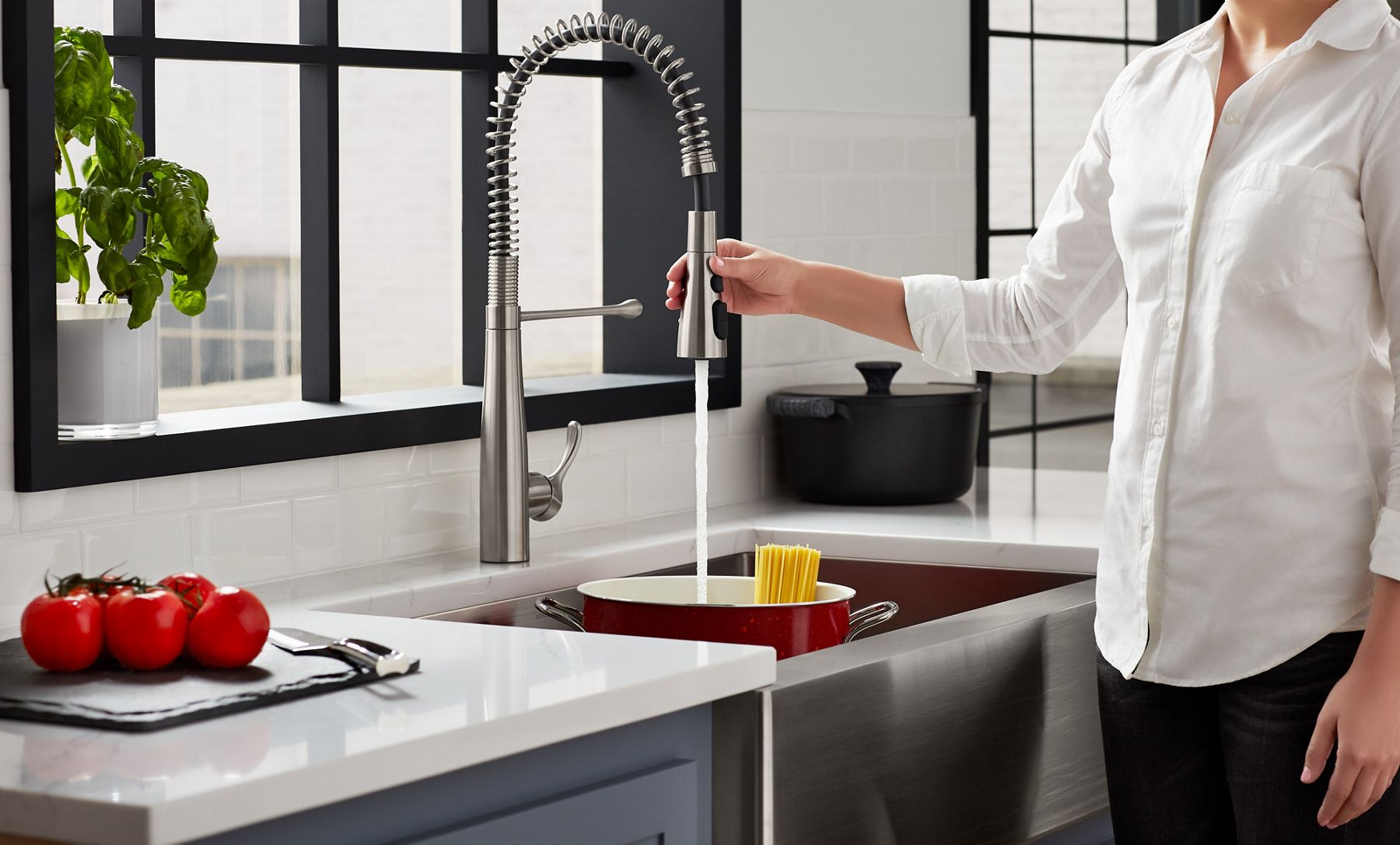 A hand holding the spout of a semi-professional faucet, pushing the Boost Spray button to fill a red enameled pot in a sink with spaghetti noodles sticking out of the top.
