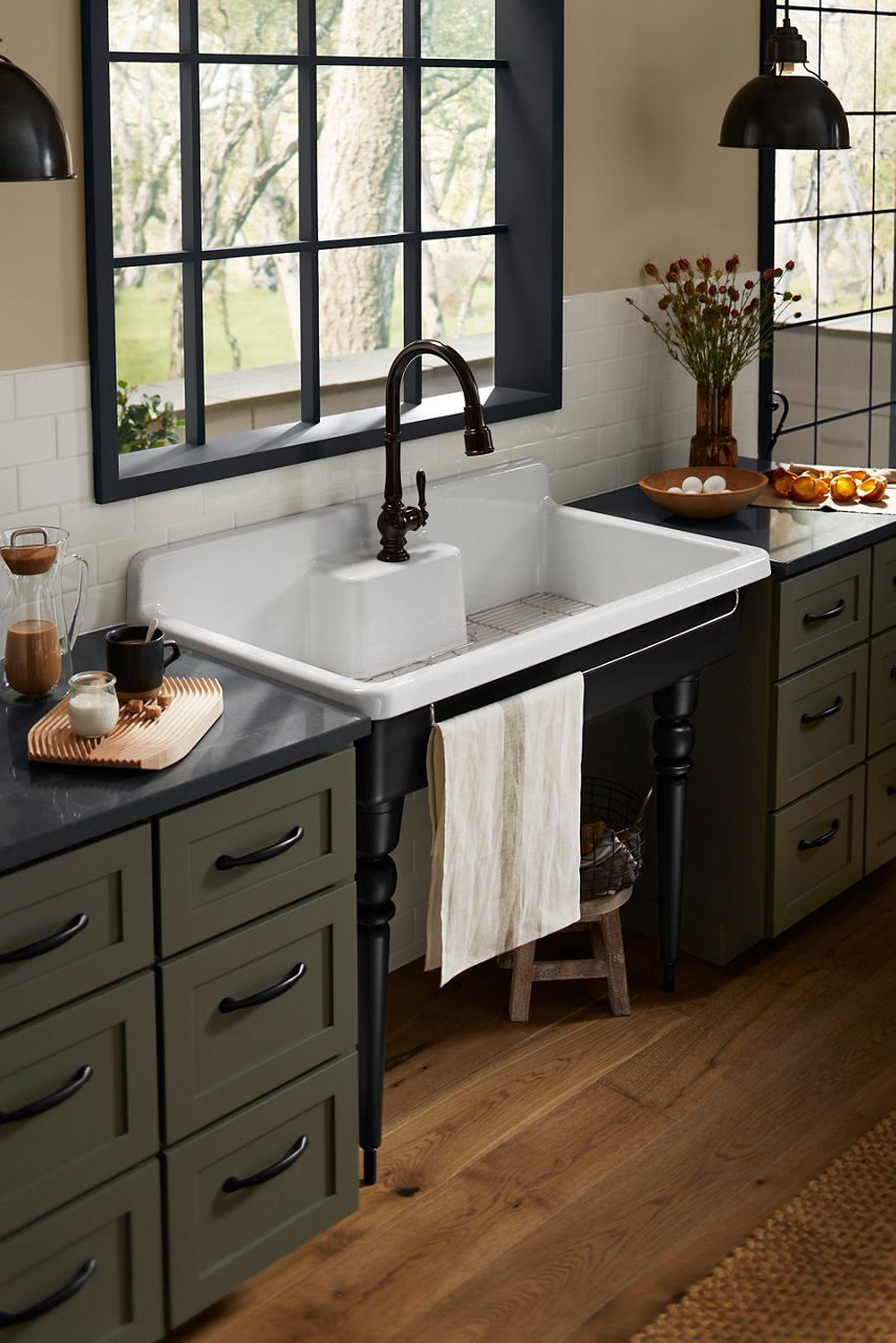 White farmhouse sink with black faucet in rustic kitchen with green cabinetry.