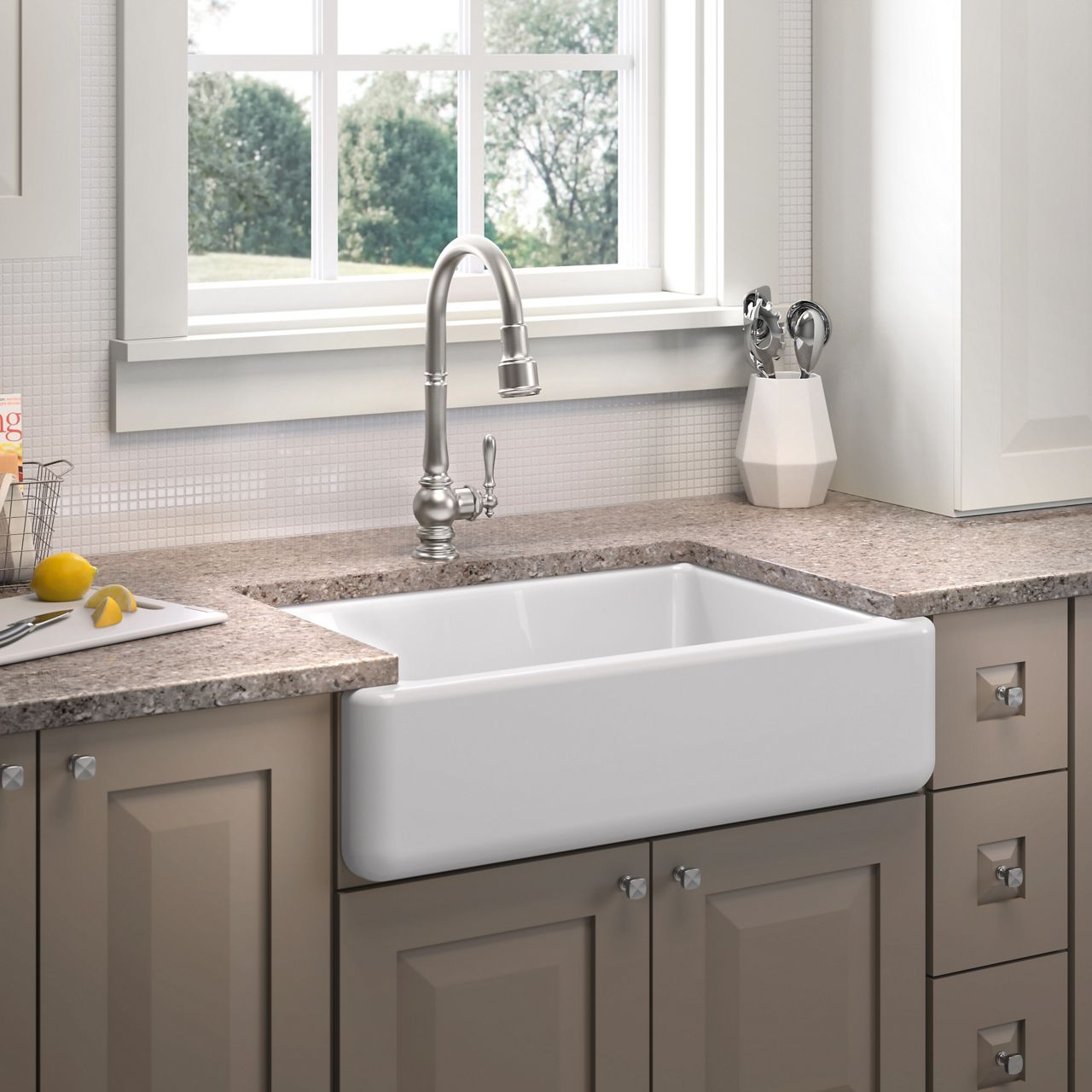 A Whitehaven apron-front sink in white, sitting in a dark tan colored cabinet with matching stone counter top under a window looking out onto a view of trees and shrubs.