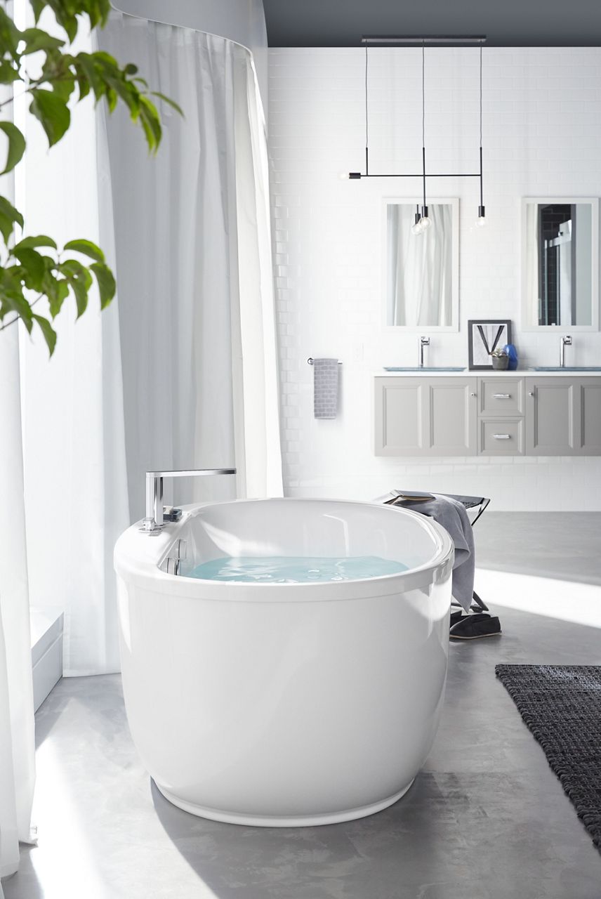 A white Sunstruck freestanding bath with a polished chrome deckmount bath faucet in a white room with bright light coming in from a nearby window.