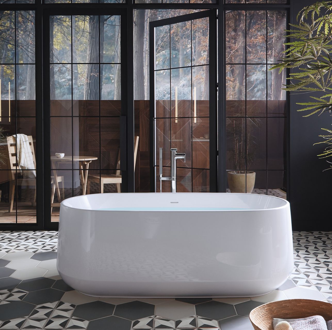 A white KOHLER Ceric freestanding bath with Composed floor mount bath filler stands before glass doors leading onto a balcony with a table and chairs.