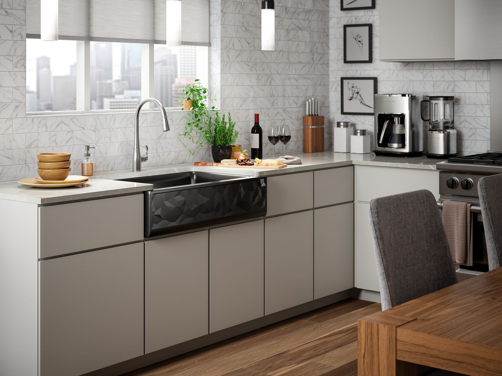 A Cairn matte black farmhouse sink with a faceted ornamental apron front set in a dark beige contemporary cabinet with a white marble tile backsplash, a stainless steel faucet and a cheese and wine display on the counter top.