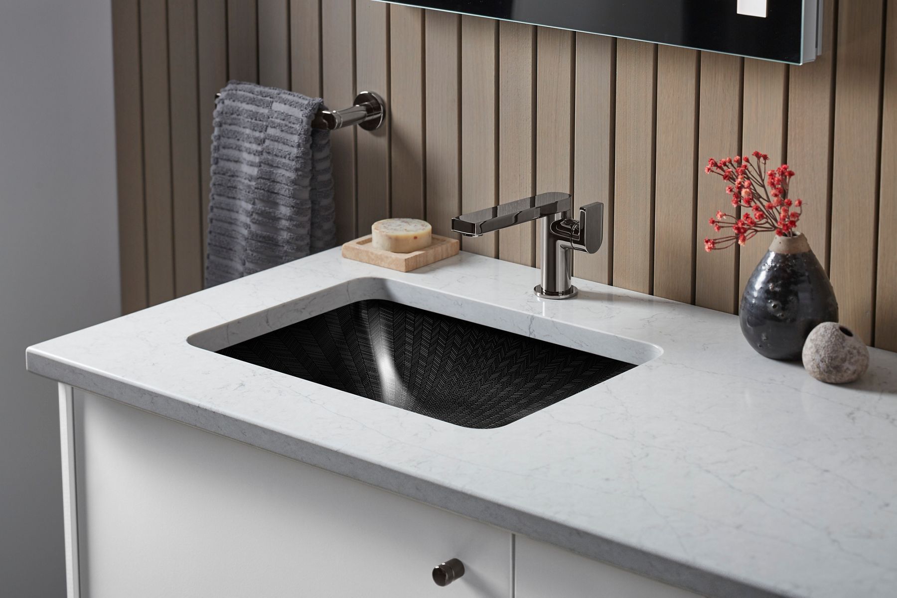 An undermount black sink with intricate herringbone detailing in a white stone counter top with a titanium finish single control faucet.