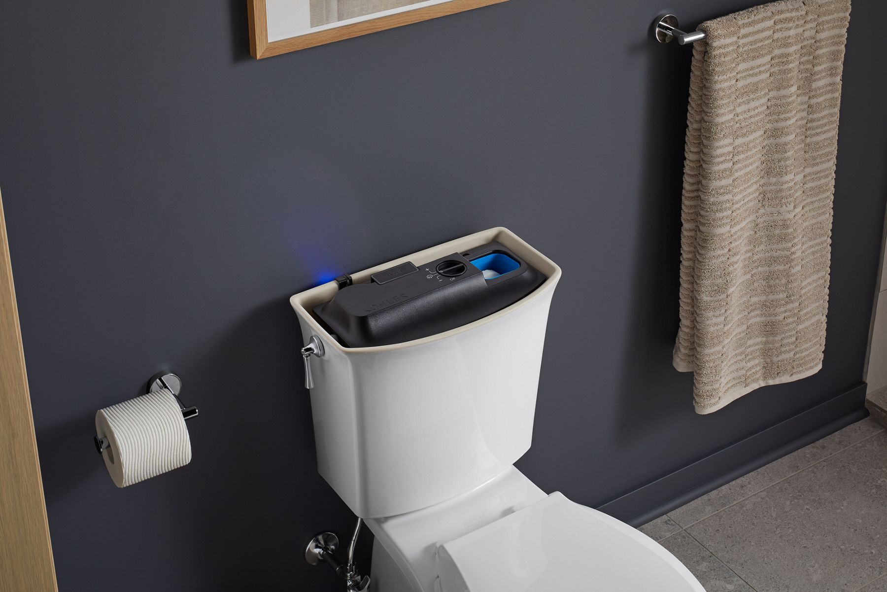 A white KOHLER toilet with tank open to reveal ContinuousClean automatic cleanser dispensing system, in a bathroom with navy blue walls and taupe curtains.