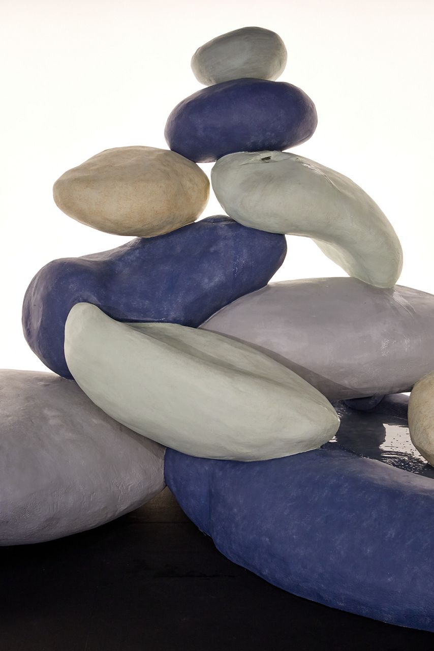 A sculpture of large violet, green, gray, and off-white stone-like objects arranged in a pile, approximately 10 objects and the sculpture is roughly 8 feet tall.