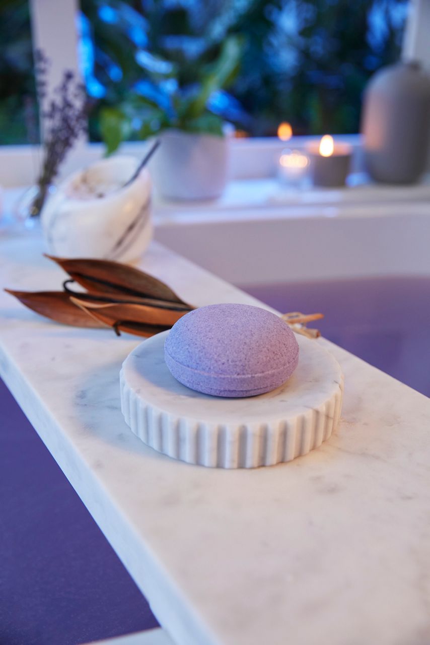 Une bombe pour le bain à la lavande placée sur un sous-verre rond et nervuré en marbre placé sur une surface en marbre lisse. À l’arrière, une bougie, une plante et un pot en marbre avec une cuillère ajoutent au cadre serein, semblable à un spa.