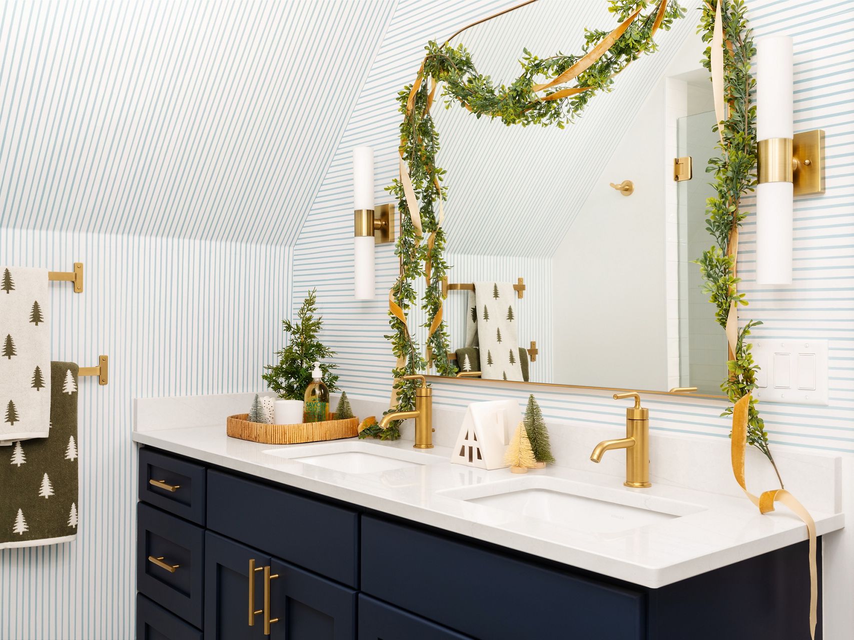 Close up image of the double-sink vanity with deep navy blue cabinet and white counter top with brass single-lever faucets, brass-trimmed mirror and brass accent wall sconce lighting. Holiday greenery with gold ribbon is draped on the mirror and counter top.