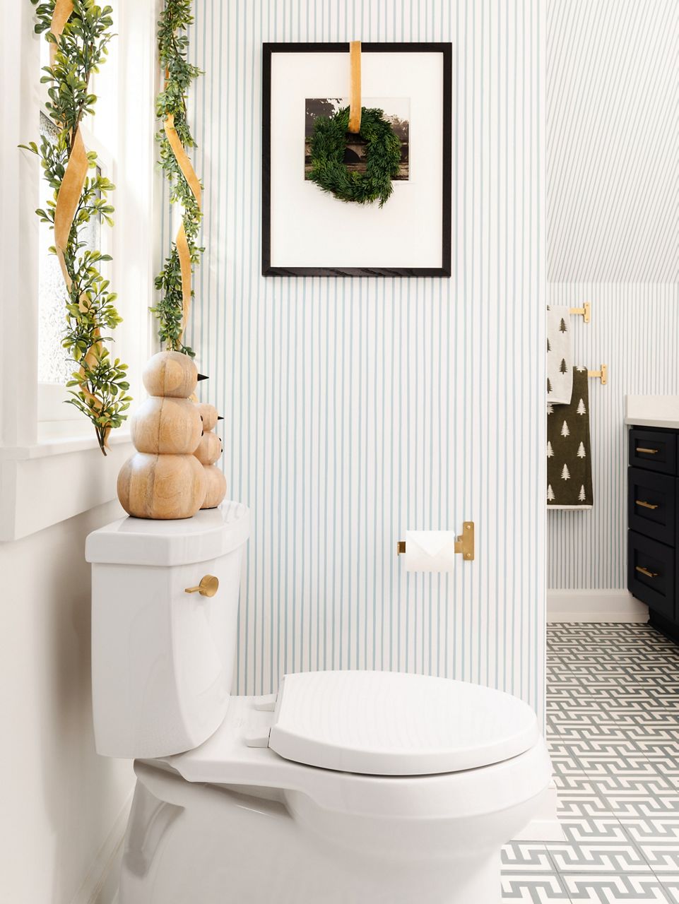 Side-profile image of the white toilet in a navy and blue and white pin-stripped wallpaper bathroom, decorated with holiday greenery and gold ribbon framing the window behind the toilet.