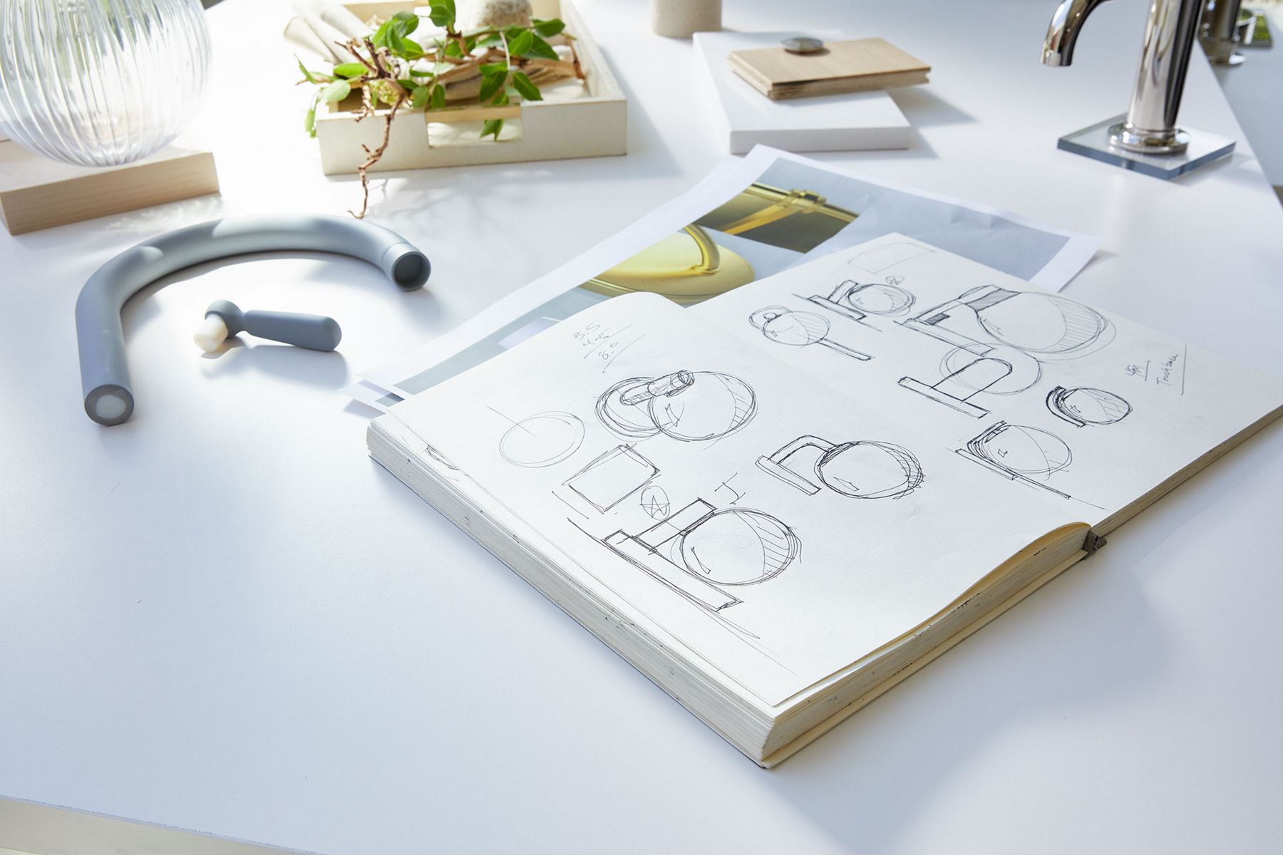 A sketchbook showing drawings of lighting that is sitting on a table next to design samples, a faucet spout and other assorted items.