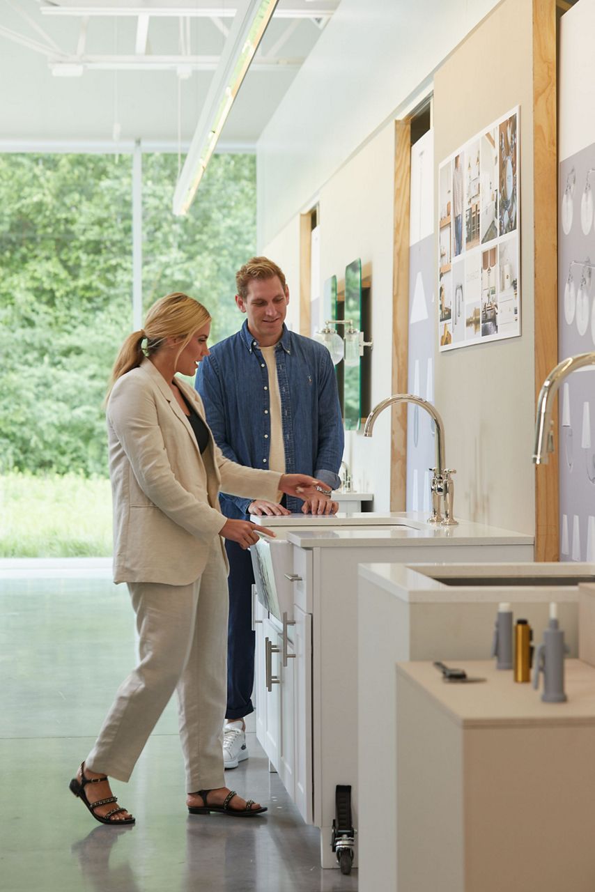Designer Shea McGee speaking with an unidentified man while looking at a kitchen cabinet, sink, and faucet display in a studio space.