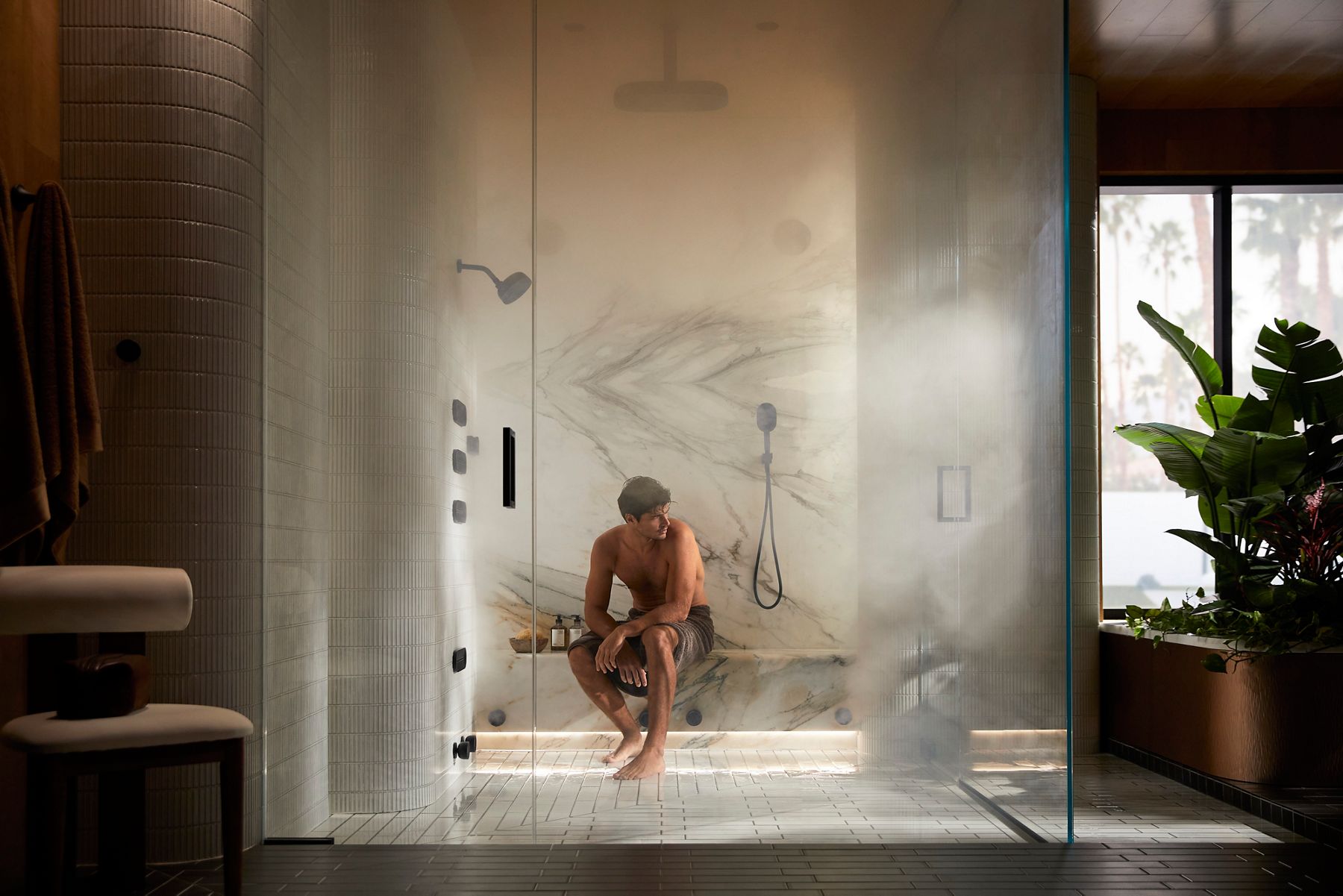 A man sitting in a glass-walled steam shower, wearing swim trunks and holding a towel.