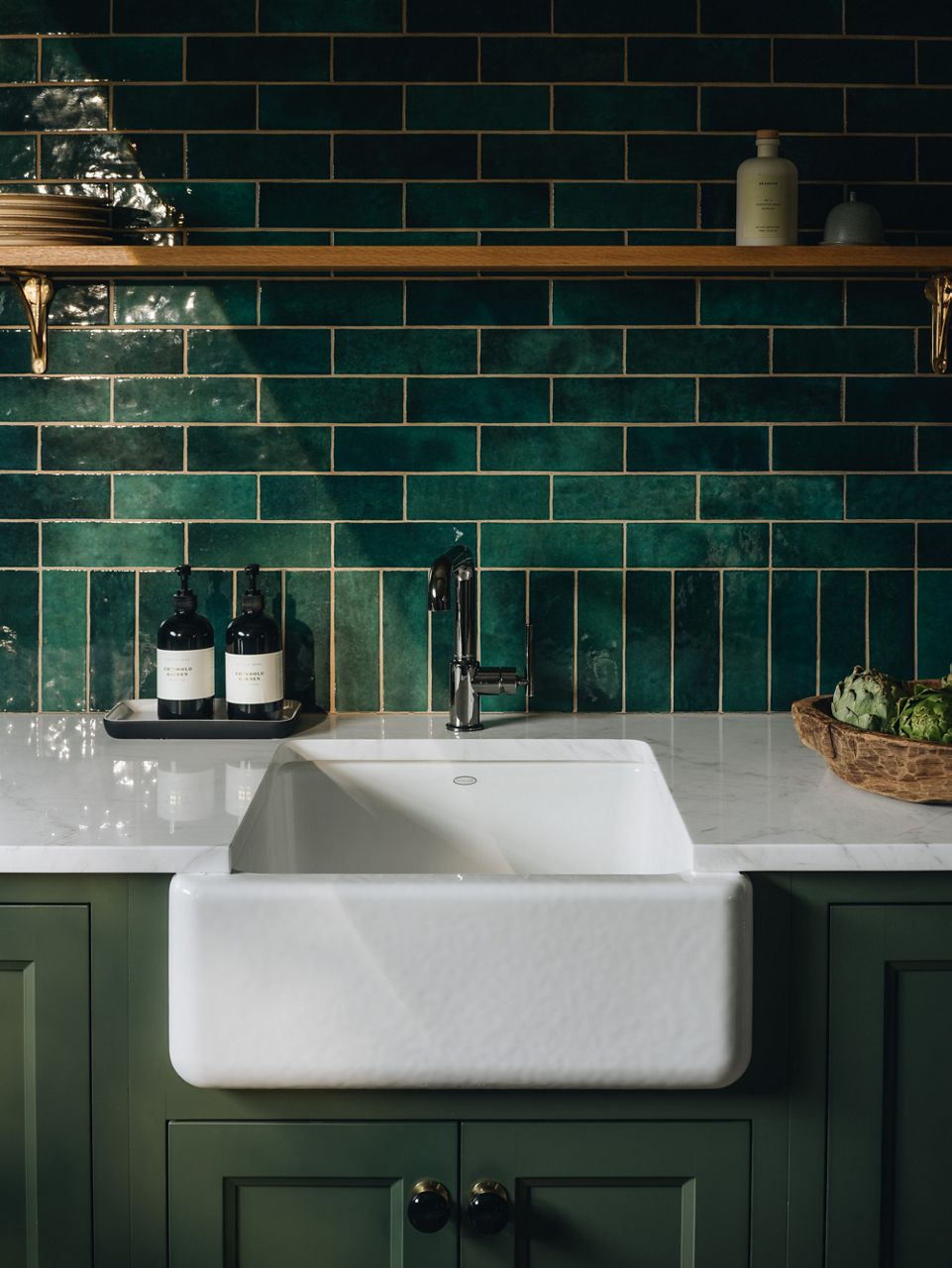 A white apron front sink installed in a kitchen with white quartz countertops  and a green tiled wall.