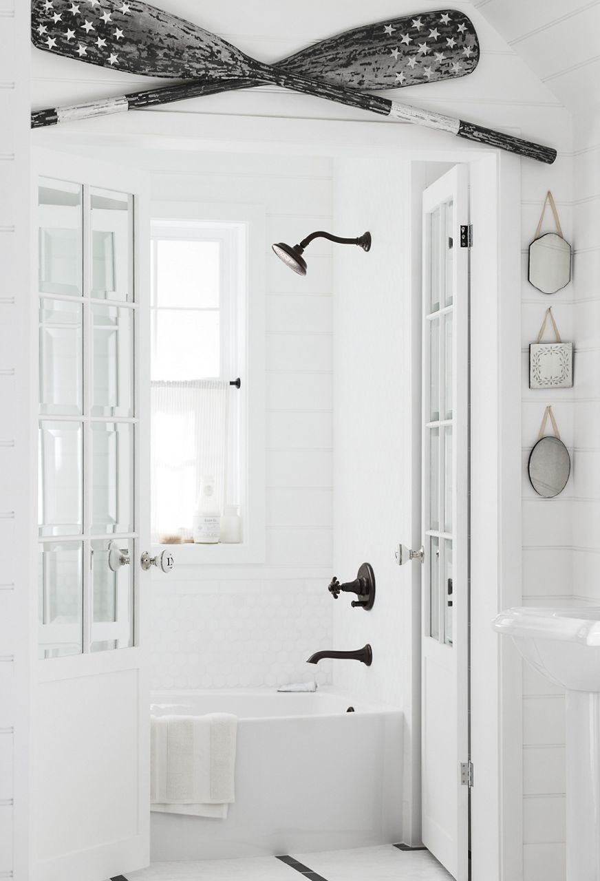 A white bathroom featuring two KOHLER pedestal sinks, black faucets with prong handles, and crossed black and white oars over the door to a separate shower enclosure.
