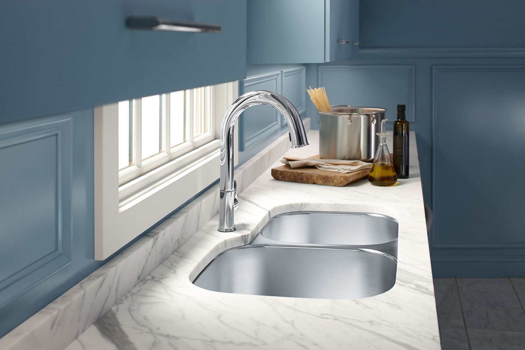 A white and gray marble kitchen counter with double bowl stainless steel sink, KOHLER touchless faucet with pullout sprayer, and a pasta pot, oil bottles, and cutting board ready for dinner prep.