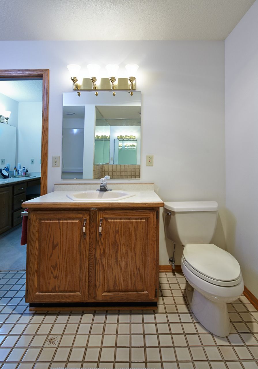 Une salle de bain désuète avec plancher en carrelage carré, meuble-lavabo au fini chêne, grand miroir et toilette allongée.