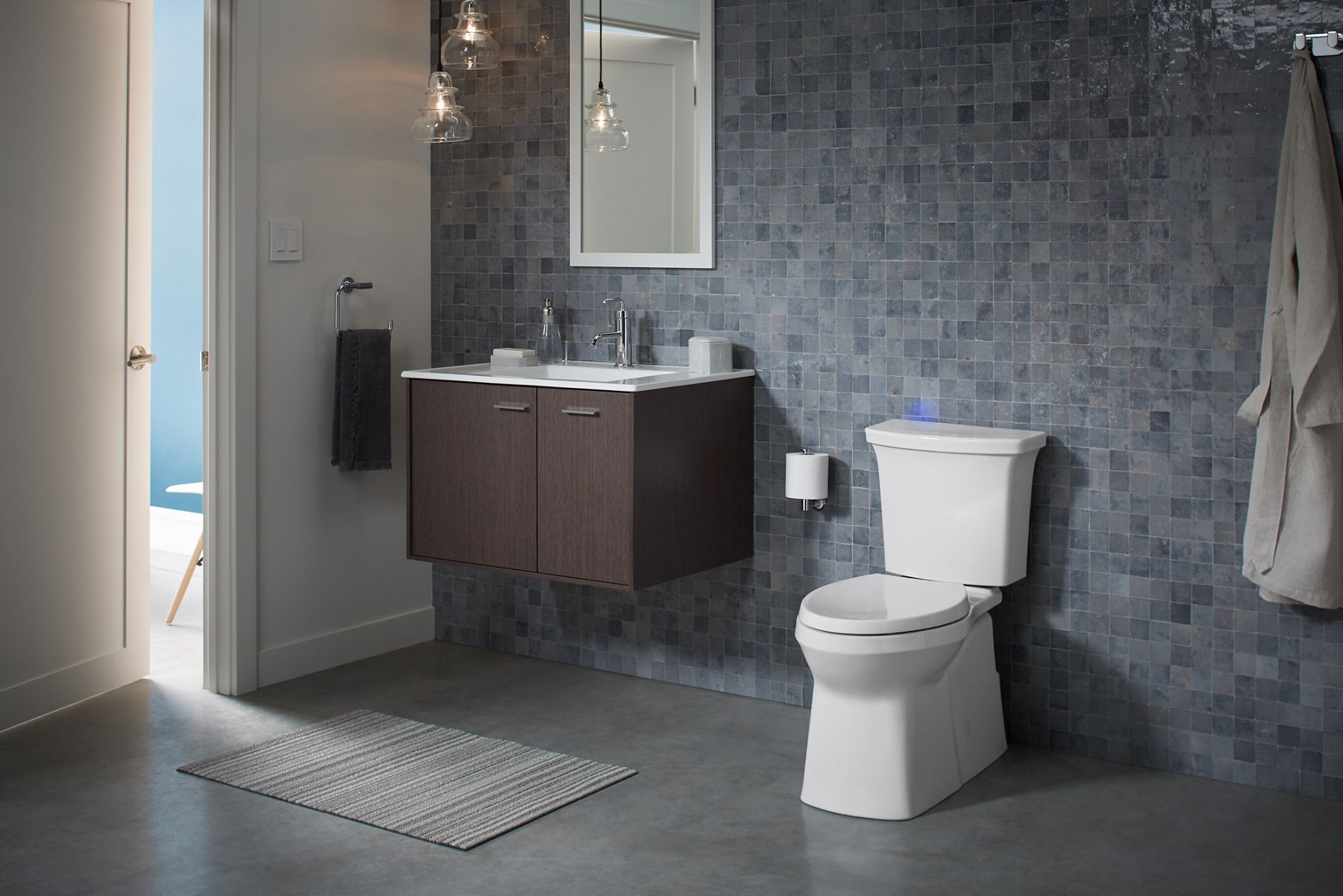 A bathroom with square blue and gray tiled wall, gray floors, wall-hung vanity, and a white KOHLER skirted trapway toilet.