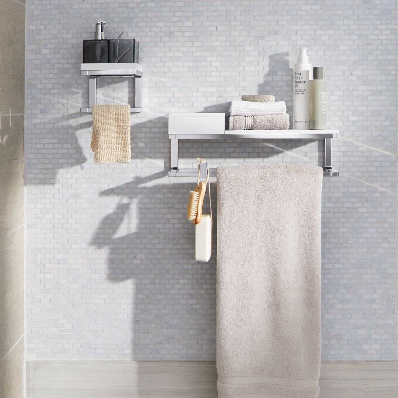 Une configuration de salle de bain moderne avec un mur carrelé gris clair. Deux tablettes en métal contiennent des serviettes bien pliées, des bouteilles, un distributeur de savon, et une brosse accrochée. Une serviette beige est pliée sur la tablette inférieure et une débarbouillette pend sur le côté, conférant à l’espace une allure organisée et minimaliste.