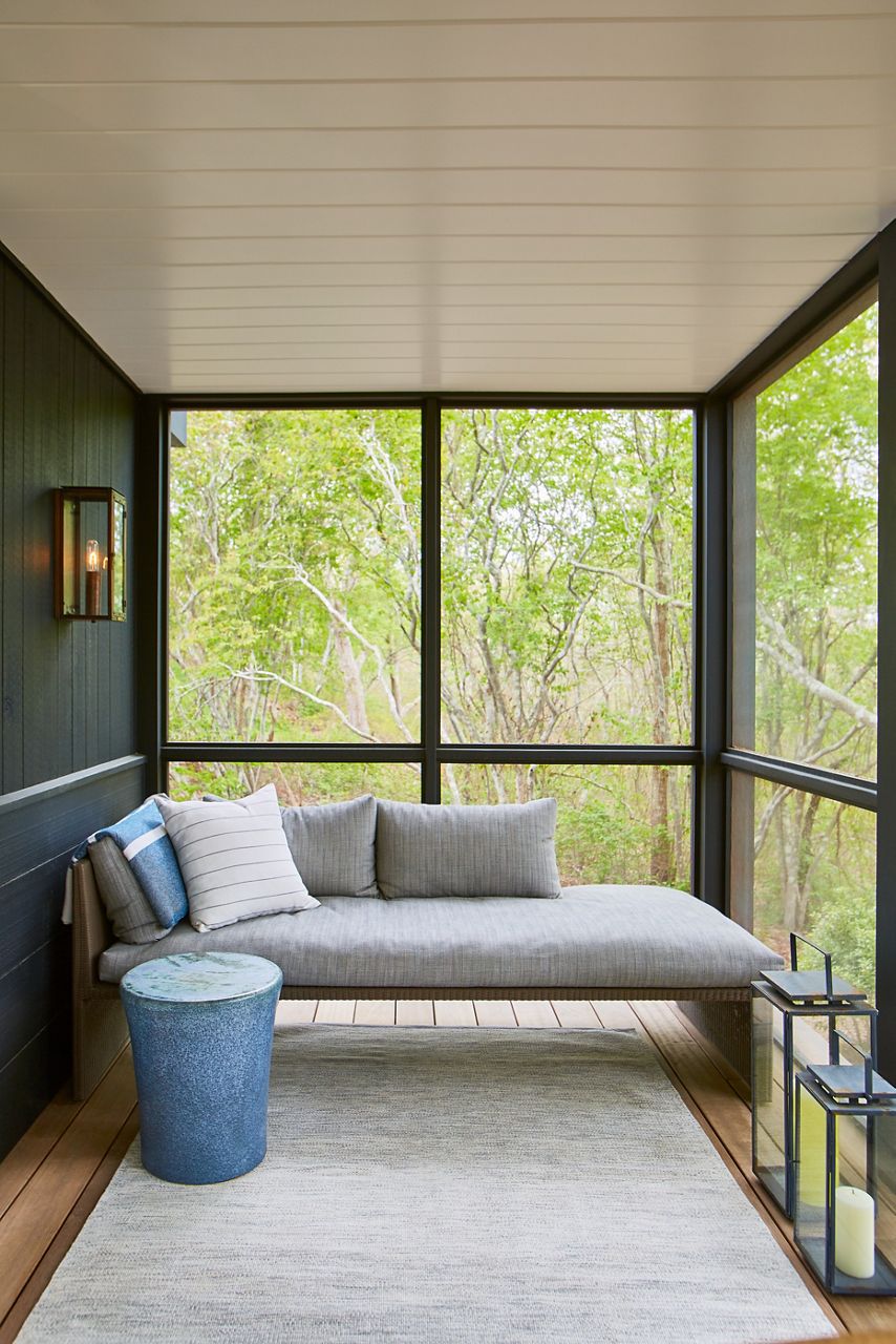 Un porche avec chaise longue rembourrée neutre, une table d’appoint ronde bleue et un tapis naturel, avec vue sur le paysage boisé.