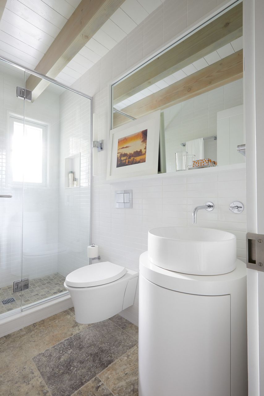Une salle de bain avec murs en carrelages blancs, plancher et plancher de douche en pierre naturelle, toilette Veil blanche au mur et meuble-lavabo rond blanc avec lavabo en surface rond Vox et robinet mural One.