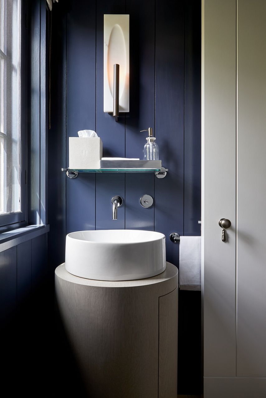 Un cabinet de toilette bleu marine avec lavabo en surface rond blanc, robinet mural, étagère pour accessoires et applique murale.